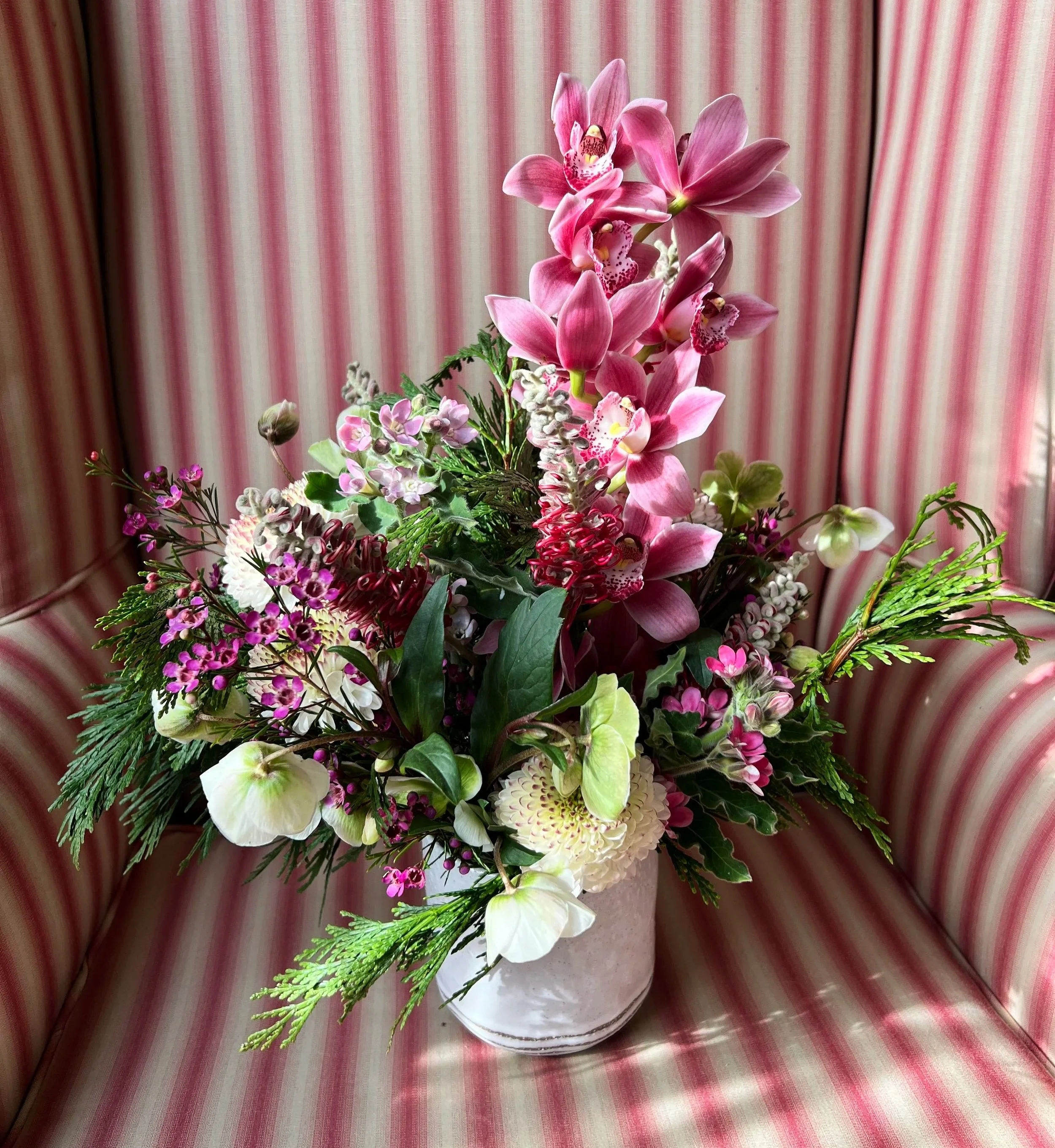 A bouquet of pink and white orchids, lilies, and greenery arranged in a glass vase on a pink and white striped fabric background.