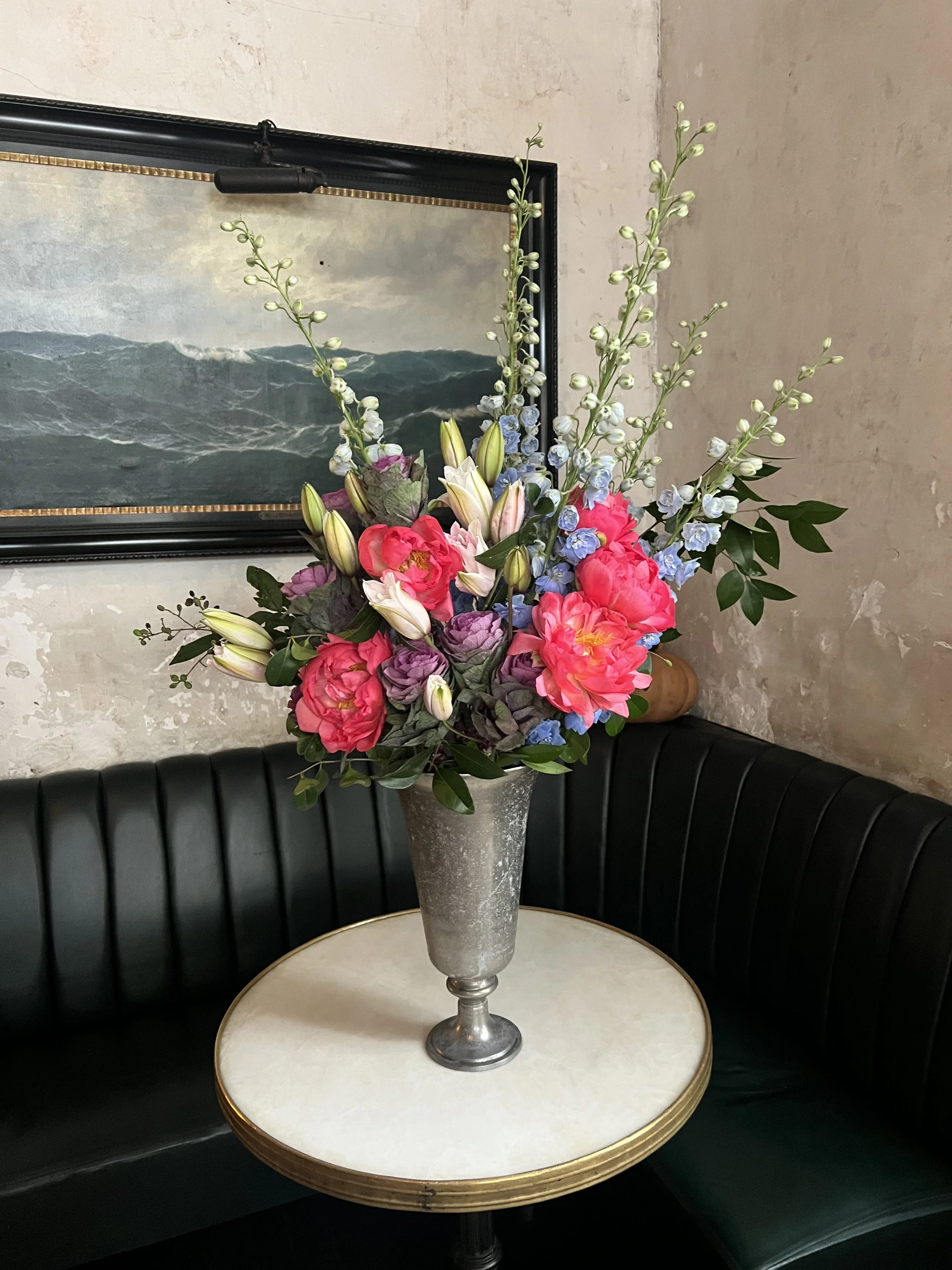 A tall marble table with a large vase filled with colorful flowers, sitting between two black curved booths against a plain textured wall.