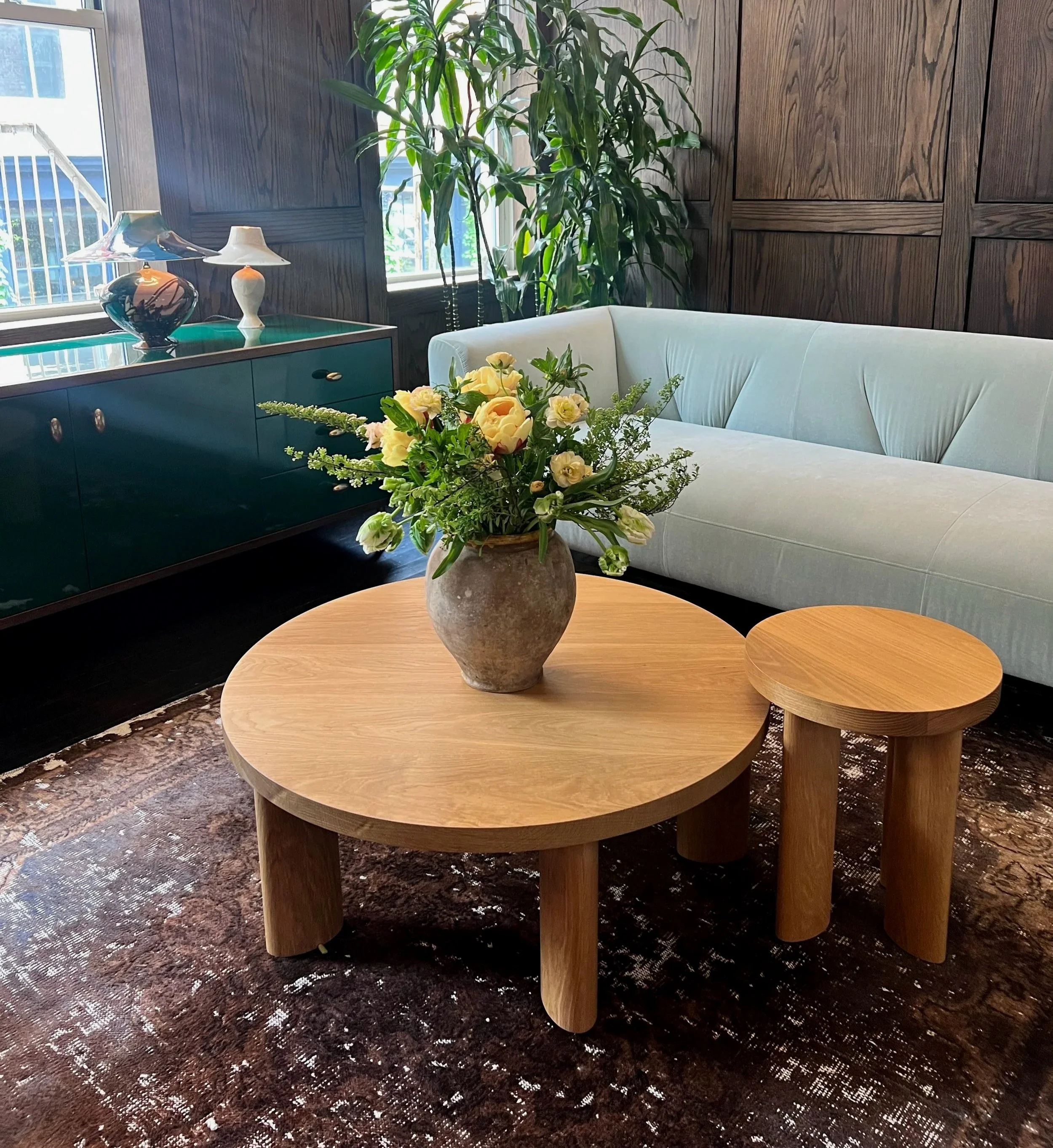 A living room with a large light gray sofa, a round wooden coffee table, a smaller matching side table, a black and brown patterned carpet, a dark wood-paneled wall, a tall indoor plant near a window, a dark green cabinet with ceramic table lamps and decorative vases, and a floral arrangement in a stone vase on the coffee table.