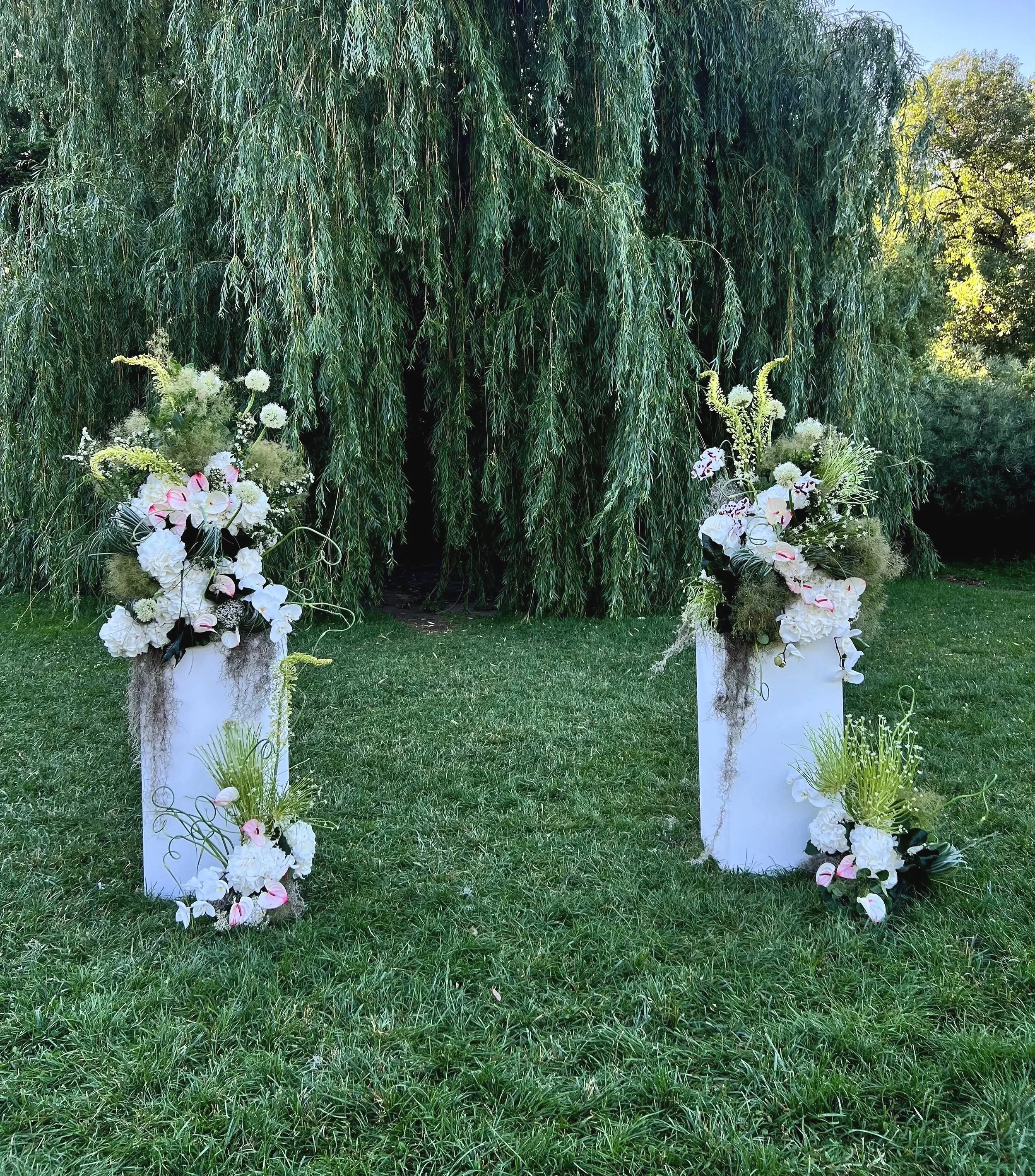 Decorative floral arrangement with white flowers and greenery, placed on white pedestals in front of a large green leafy tree on a grassy lawn.