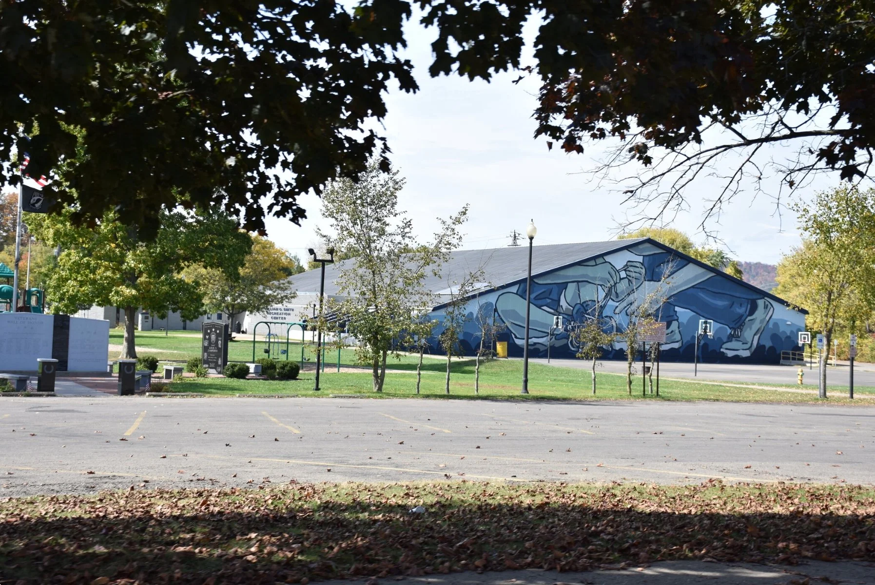 A park with a large mural of a dog painted on the side of a building. The park has trees, a playground, some signs, and a mostly empty parking lot in the foreground.