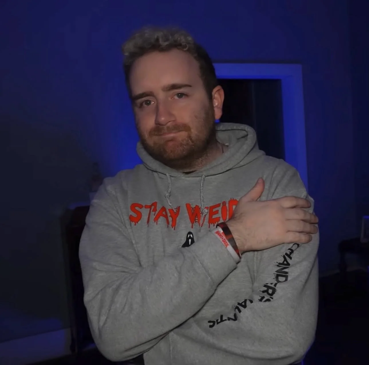 A man with light brown, curly hair and a beard, wearing a gray hoodie that says "Stay Weird" and "Chandler," standing in a dimly lit room with blue and purple lighting. He is crossing his arms over his chest, with one hand resting on his shoulder.