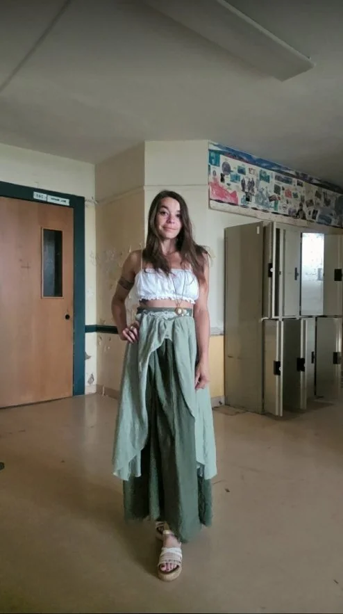 A woman with long brown hair sitting on the floor in front of lockers, smiling at the camera.