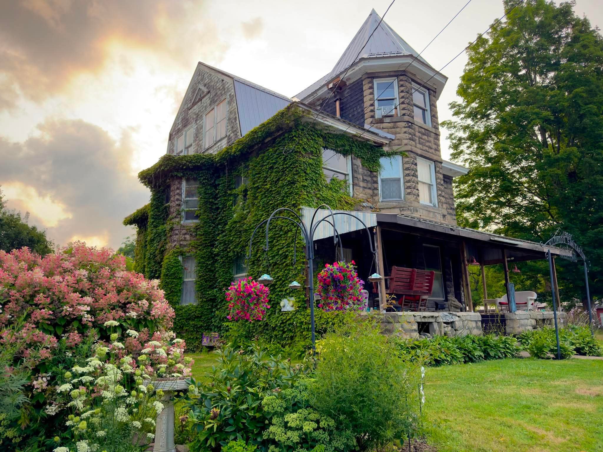 A large Victorian-style house with a stone exterior, steeply pitched roof, and numerous windows, partially covered with green ivy. There is a garden with colorful flowers and a lush green lawn in front, and a large green tree to the right side. The sky above has a golden glow with some clouds.
