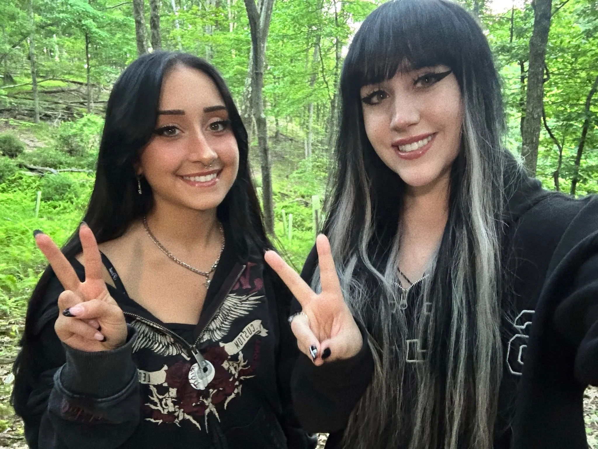 Two women outdoors in a forest, smiling and making peace signs with their fingers.