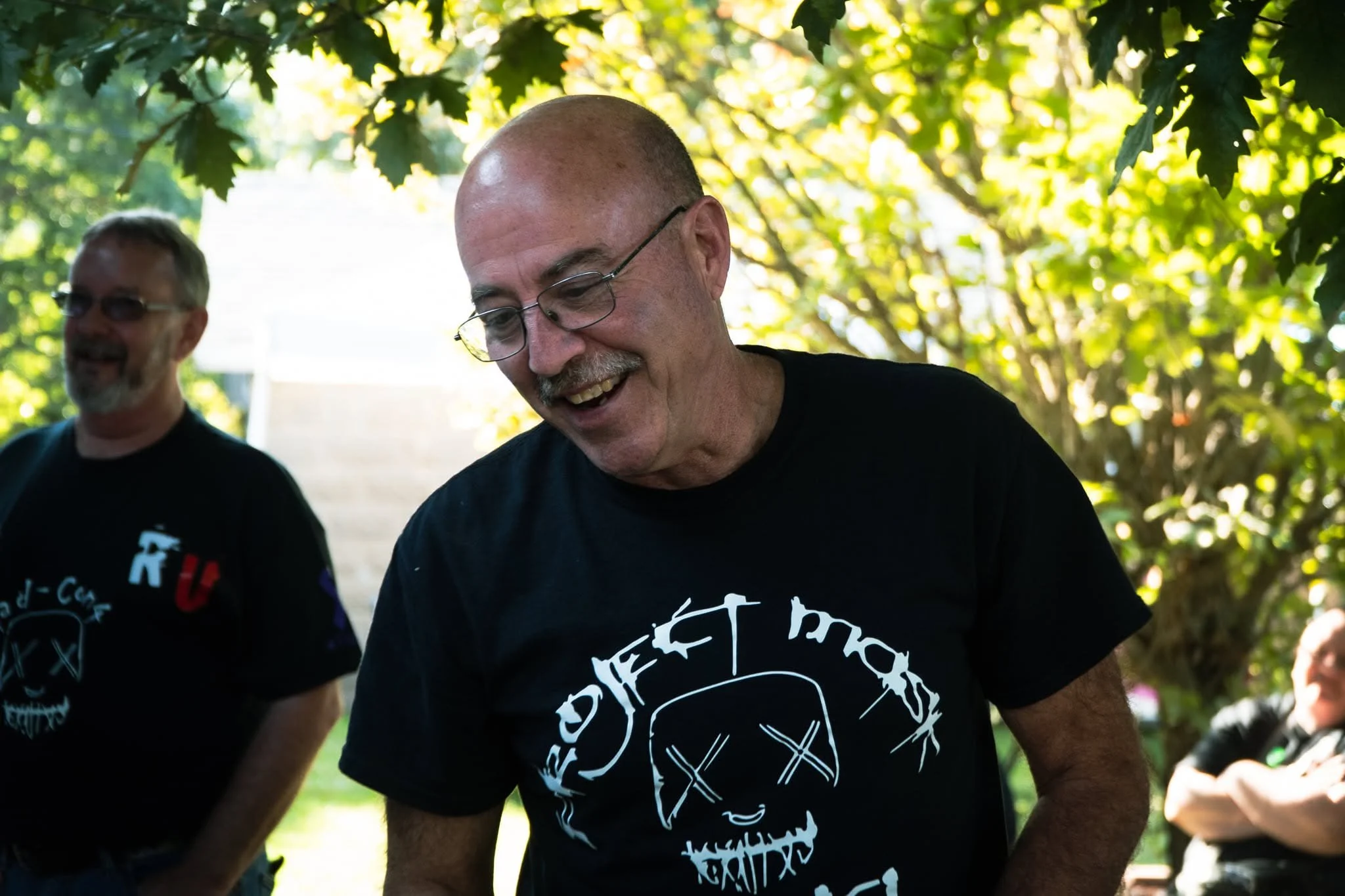 Two men outdoors under trees, smiling and laughing. One man has a shaved head, glasses, and a mustache. The other man has gray hair, sunglasses, and a beard. They are wearing black t-shirts with graphic designs.