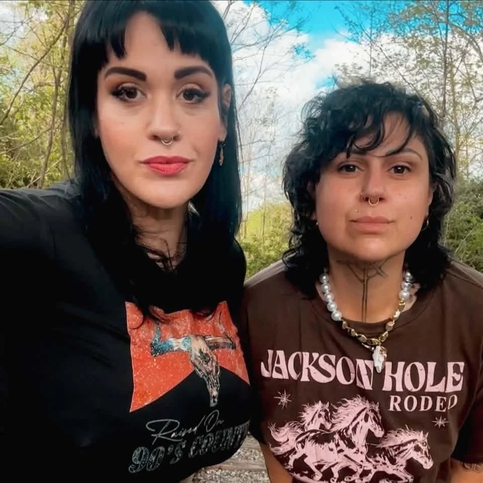 Two women standing outdoors in front of trees, both with dark hair and piercings. The woman on the left has straight black hair, makeup, and a septum piercing, wearing a graphic t-shirt. The woman on the right has curly dark hair, multiple facial piercings, and tattoos, wearing a brown t-shirt with horses and text, along with jewelry.