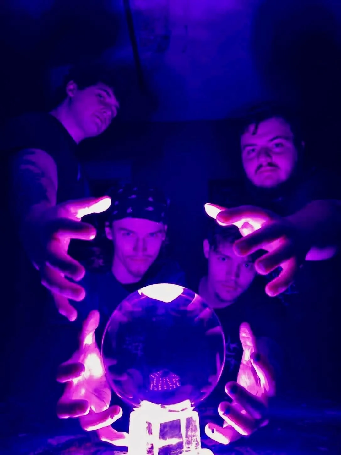 A group of four young men in a dark room with purple lighting, gathered around a crystal ball with a glowing base, some pointing at it.
