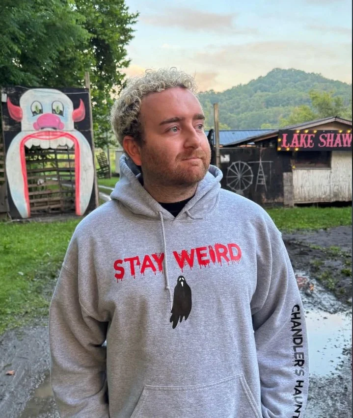 A man with light brown, curly hair and a beard, wearing a gray hoodie that says "Stay Weird" and "Chandler," standing in a dimly lit room with blue and purple lighting. He is crossing his arms over his chest, with one hand resting on his shoulder.