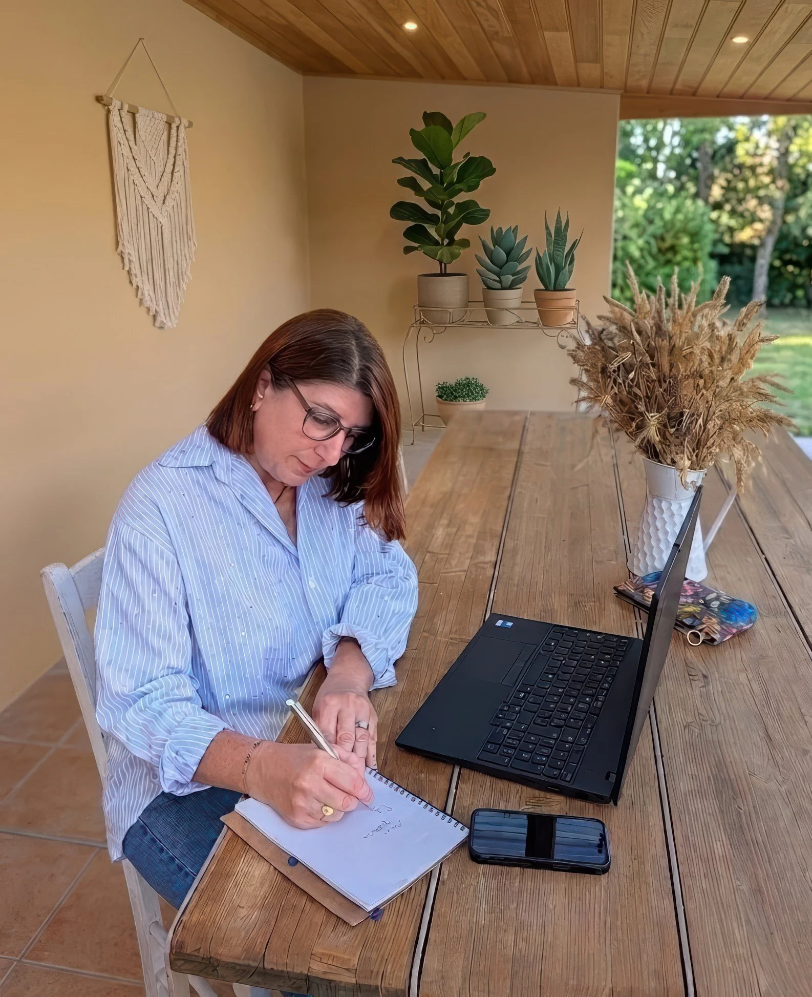 Une femme avec des cheveux bruns et des lunettes écrit dans un cahier à côté d'un ordinateur portable sur une table en bois. La scène est dans une pièce avec un mur beige, une décoration en macramé, des plantes en pot et une grande fenêtre laissant entrer la lumière naturelle.
