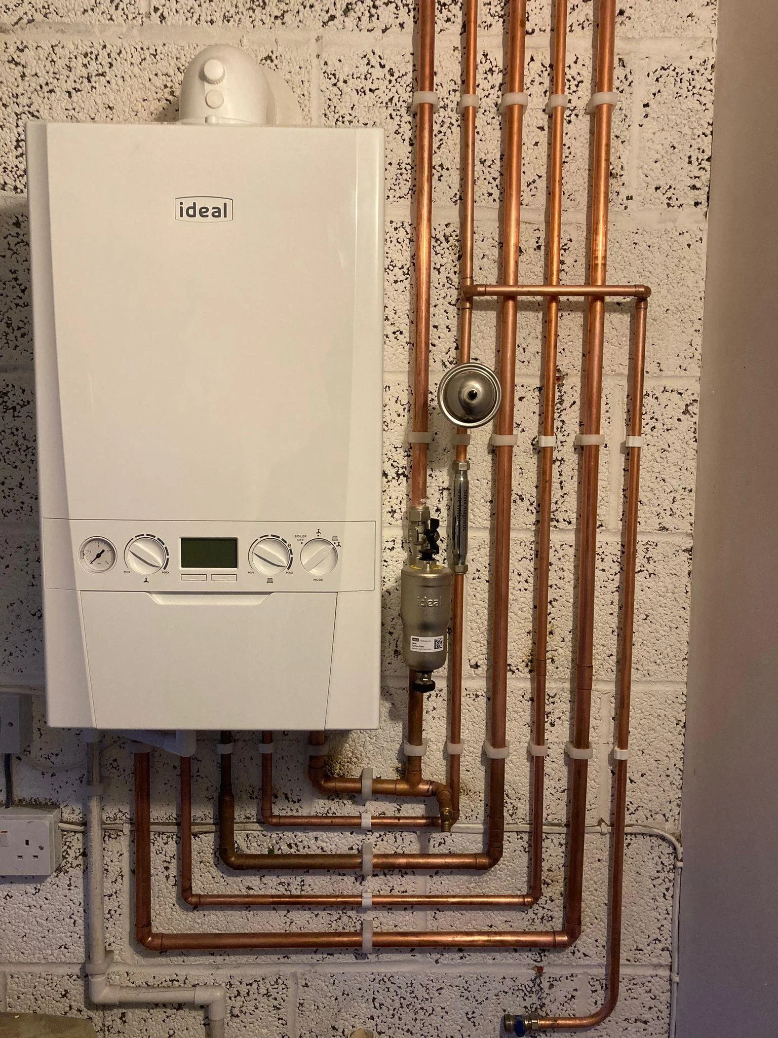 A wall-mounted boiler and copper pipes arranged vertically and horizontally, with a water filter and a power outlet nearby.