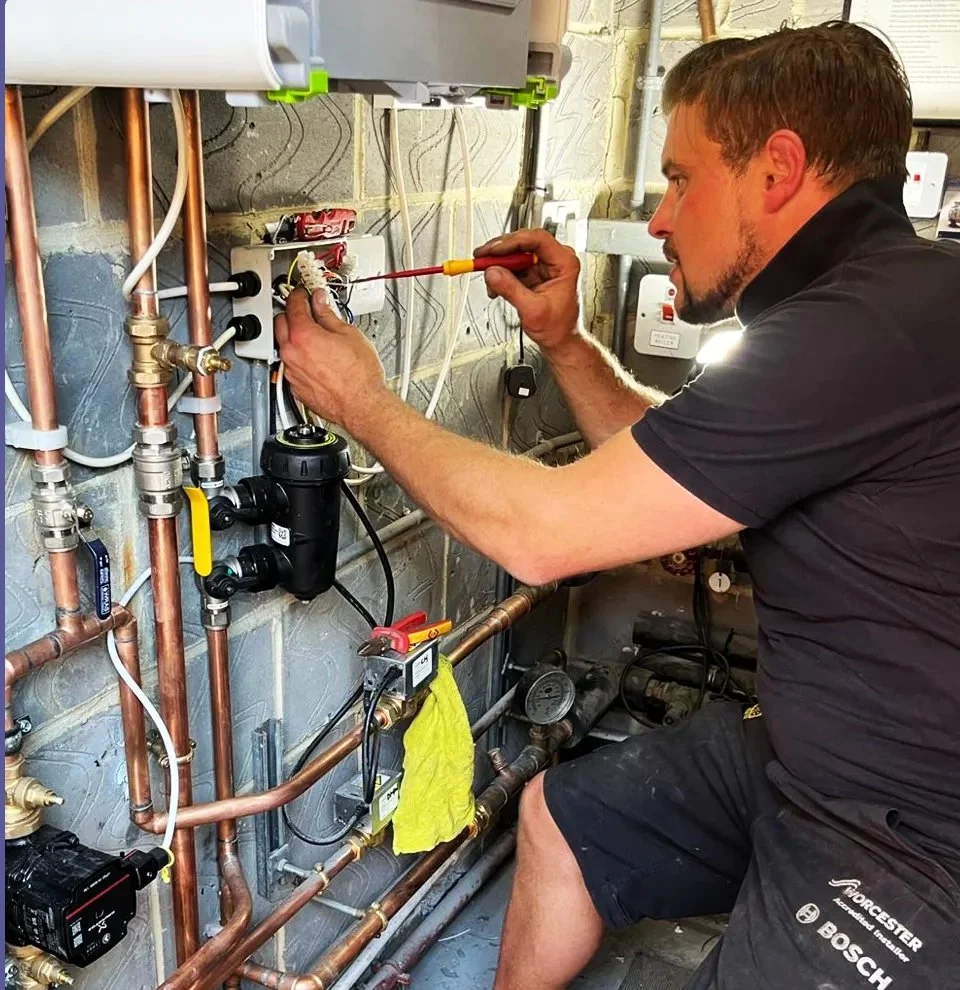 A man working on plumbing or electrical system, using a screwdriver, in a utility room with copper pipes, wires, and electrical components.