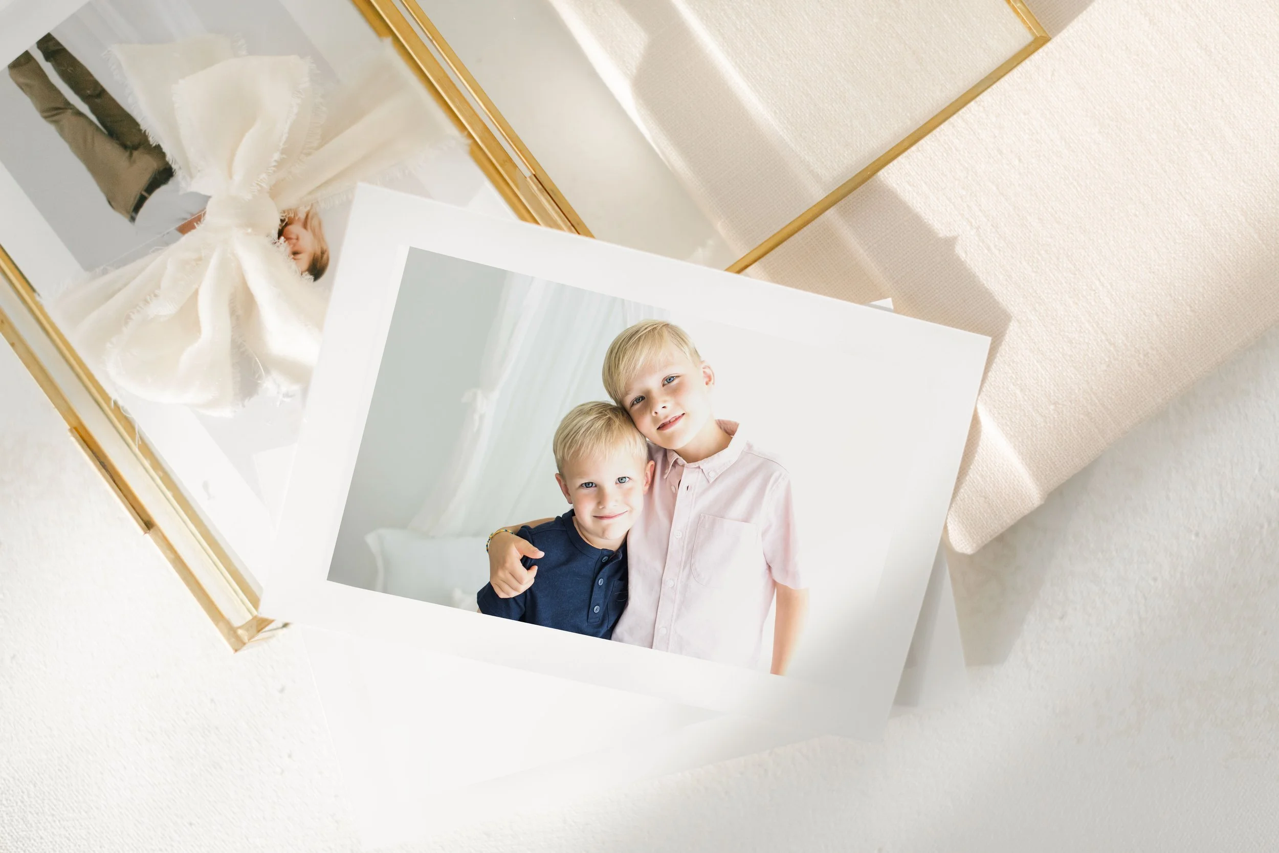 Photographs of two young boys, one with blond hair and the other with brown hair, smiling and hugging each other. The photographs are resting on a textured, light-colored surface.