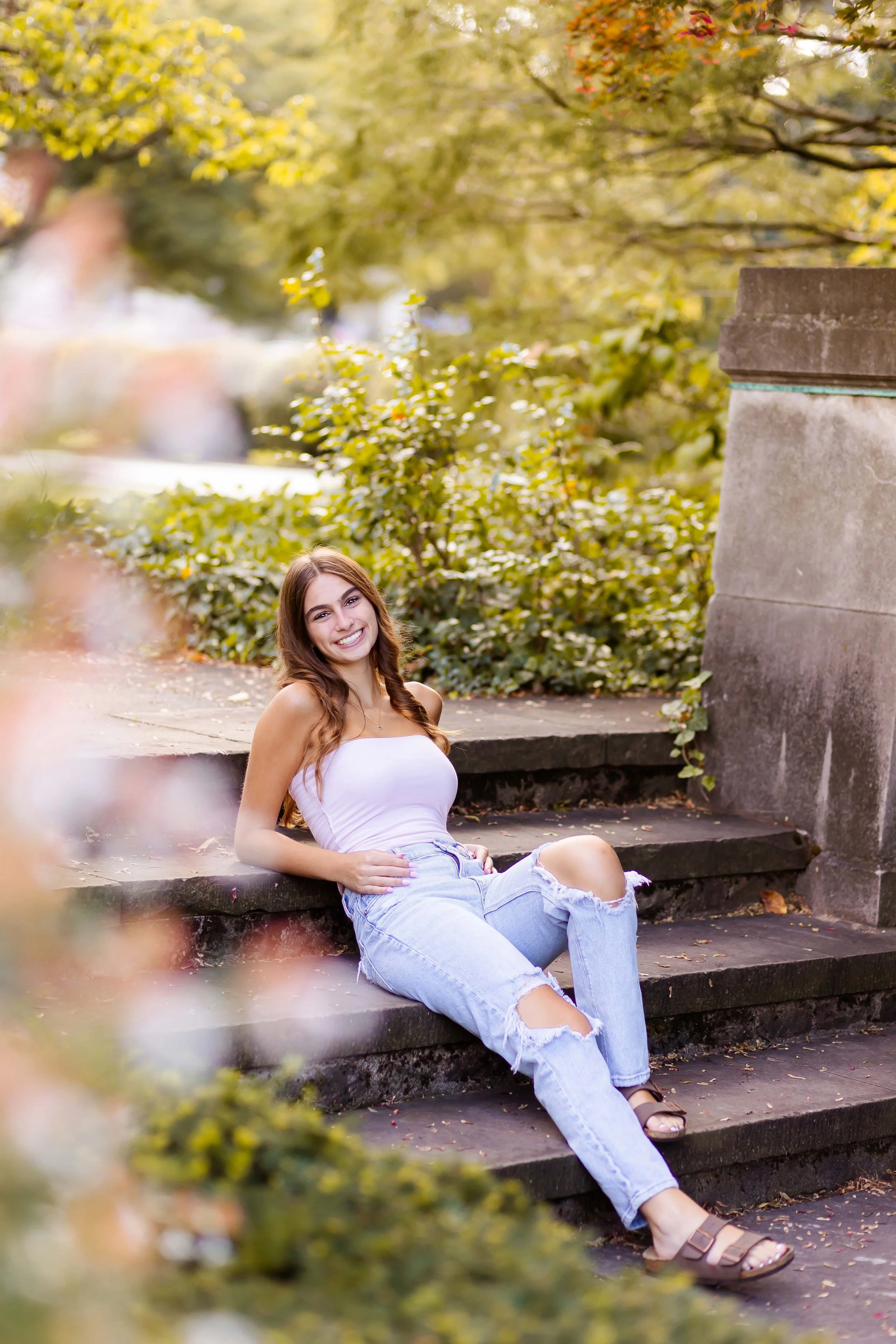 A senior girl sitting on outdoor steps, dressed in a white strapless top and ripped light blue jeans, with greenery and trees in the background during her senior portrait session with East and Elm Portrait Studio in Rochester, NY.