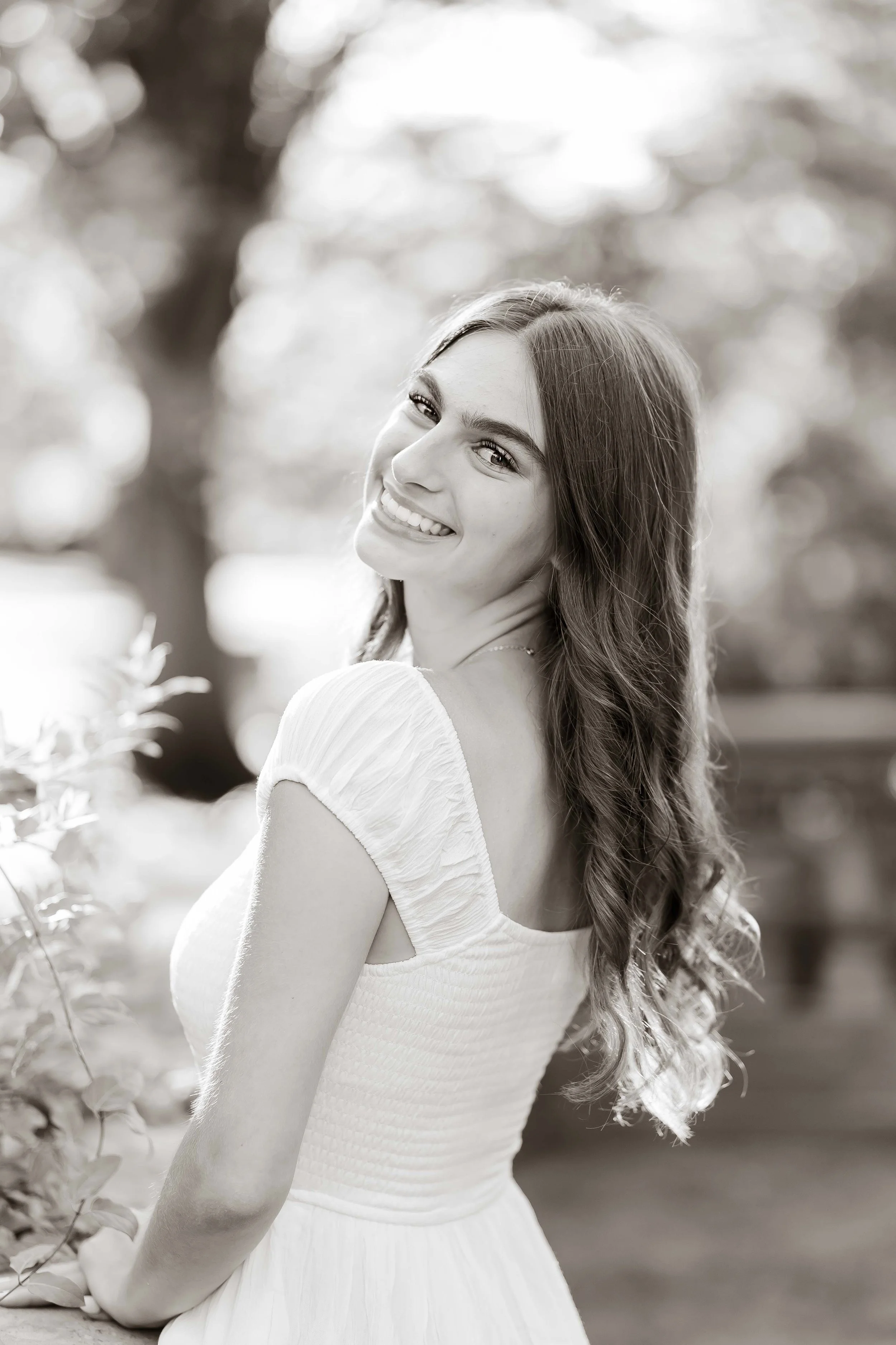 A young woman with long wavy hair smiling outdoors in a white dress during her senior portrait session with East and Elm portrait studio in Rochester, NY