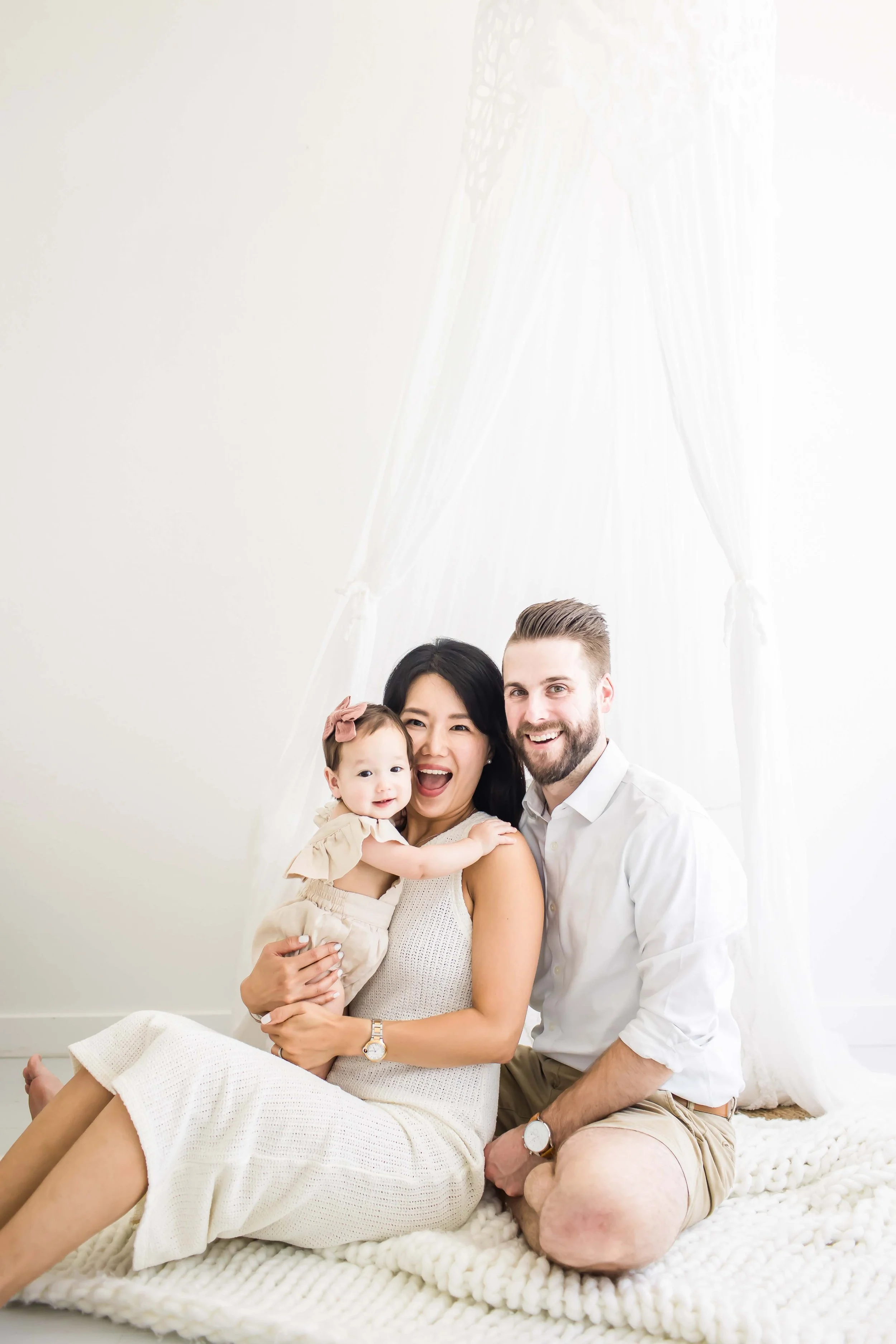 A happy family of three sitting on a cozy white blanket at East and Elm Portrait studio in Fairport, NY, during family session.