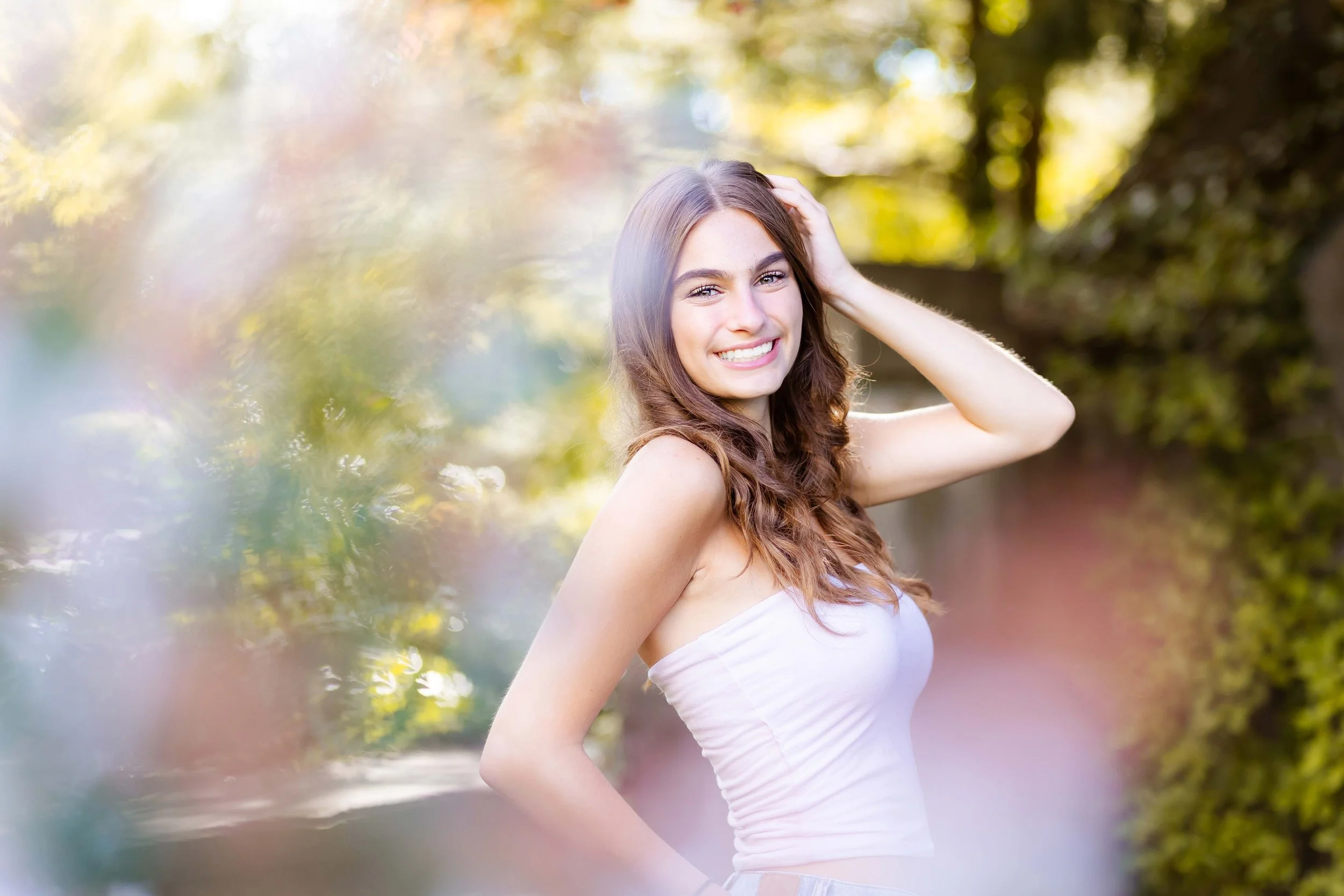 A young woman with long, wavy brown hair smiling outdoors during the daytime, with one hand touching her head, standing against a backdrop of blurred trees and sunlight during senior portrait photos with East and Elm Portrait Studio in Rochester, NY.