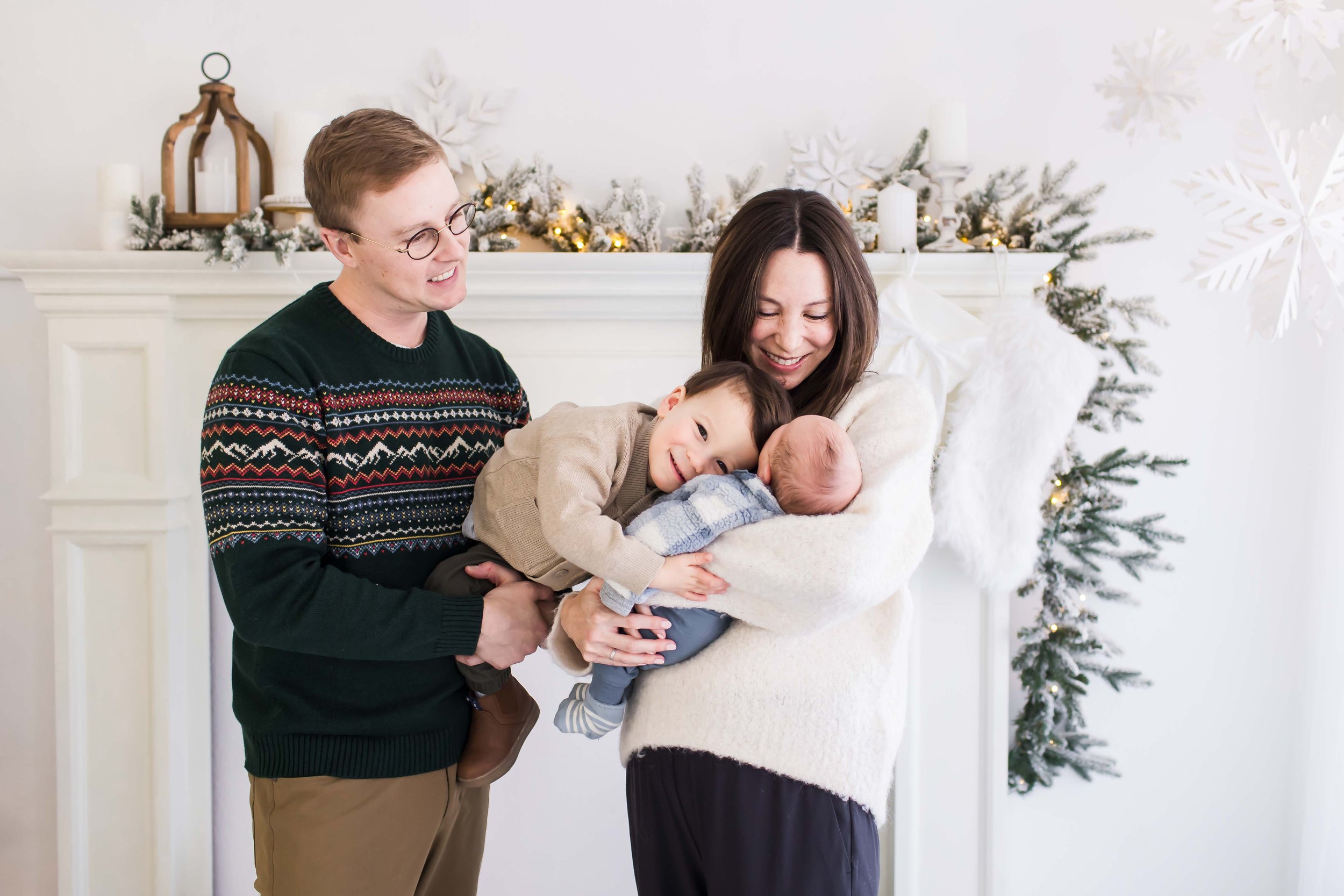 A family of four in festive attire gathered by a white fireplace with Christmas greenery and lights, celebrating Christmas at East and Elm Portrait Studio in Fairport, NY, during a Christmas mini session.