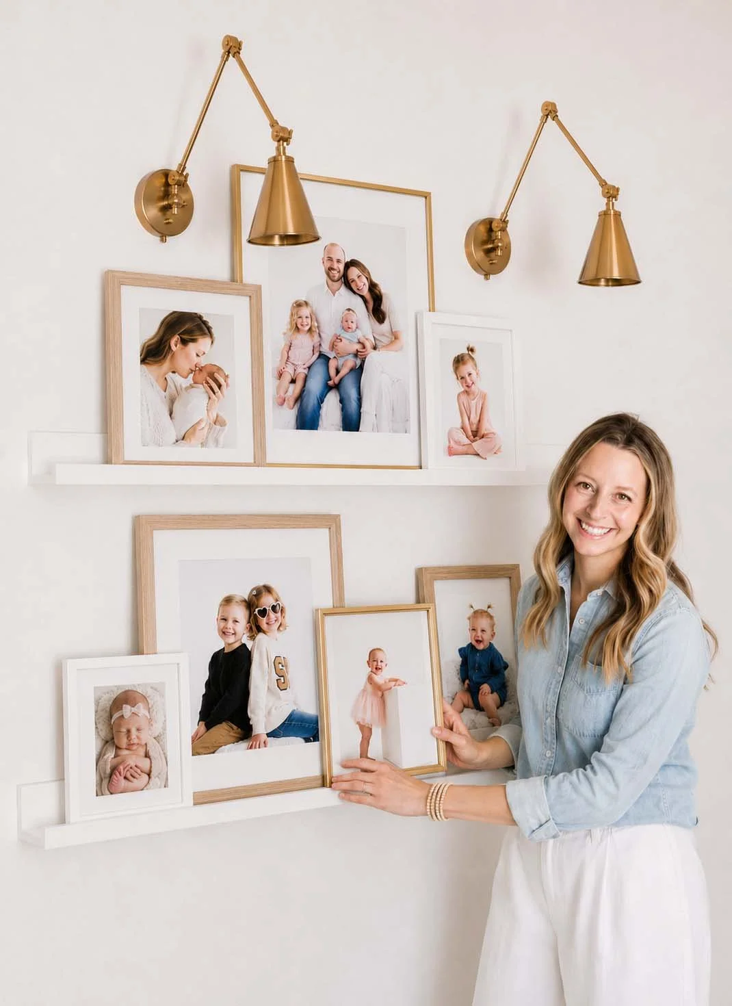 A woman with long wavy hair wearing a light blue shirt and white pants, smiling and holding a framed photo of a small girl in a pink dress. Behind her are multiple framed family photos arranged on a white wall, with gold sconces above.
