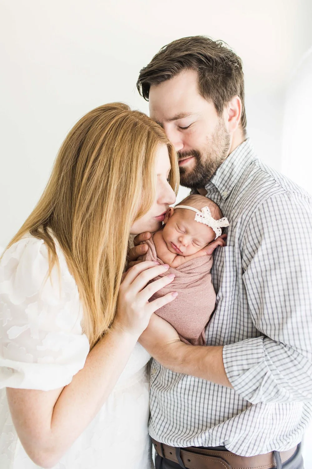 A couple holding their newborn baby, with the mother kissing the baby's forehead, and both parents smiling tenderly at East and Elm Portrait Studio during a studio newborn photography session in Fairport, NY.