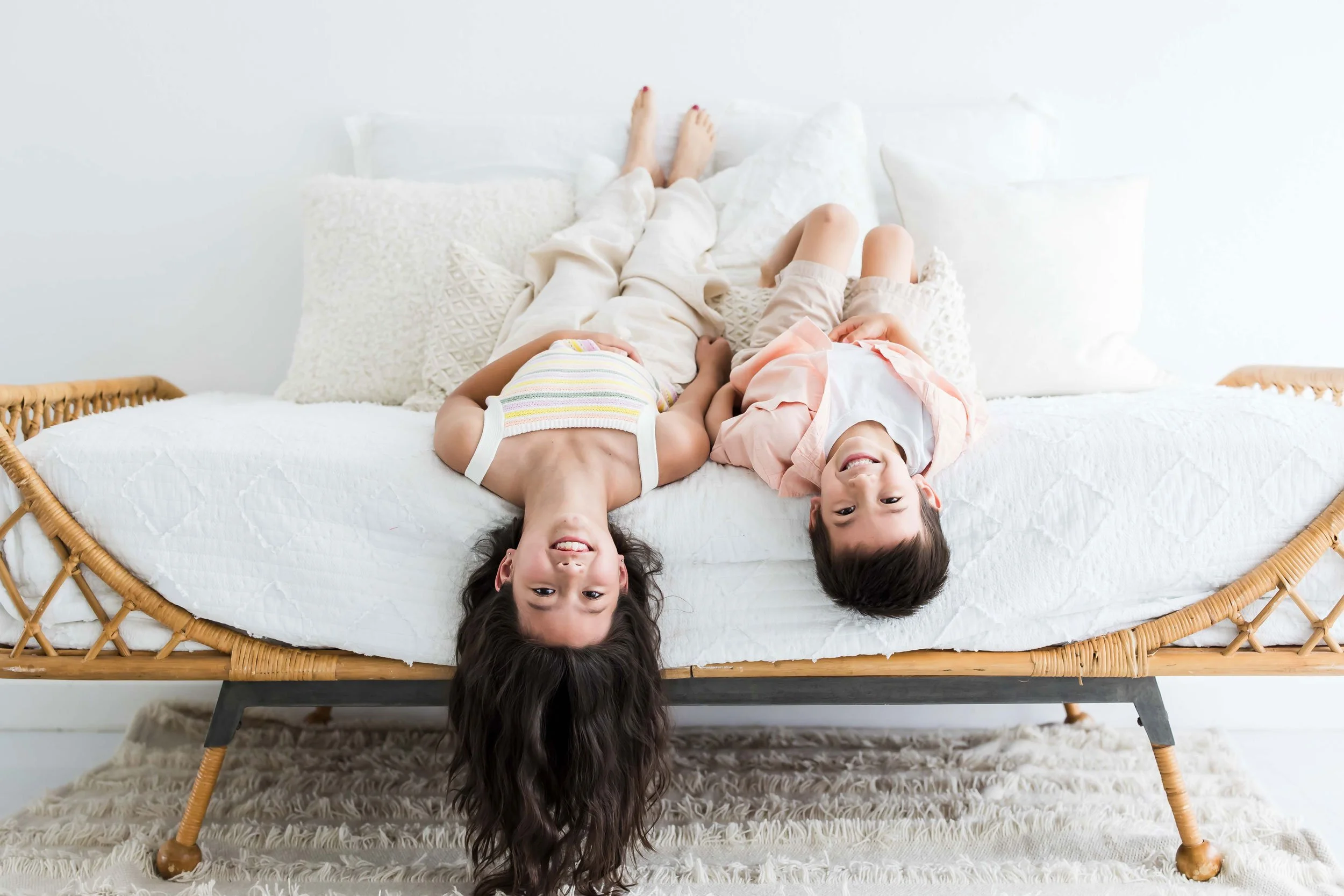 Two children lying upside down on a bed, smiling and playing at East and Elm Portrait Studio in Fairport, NY, during a family session.