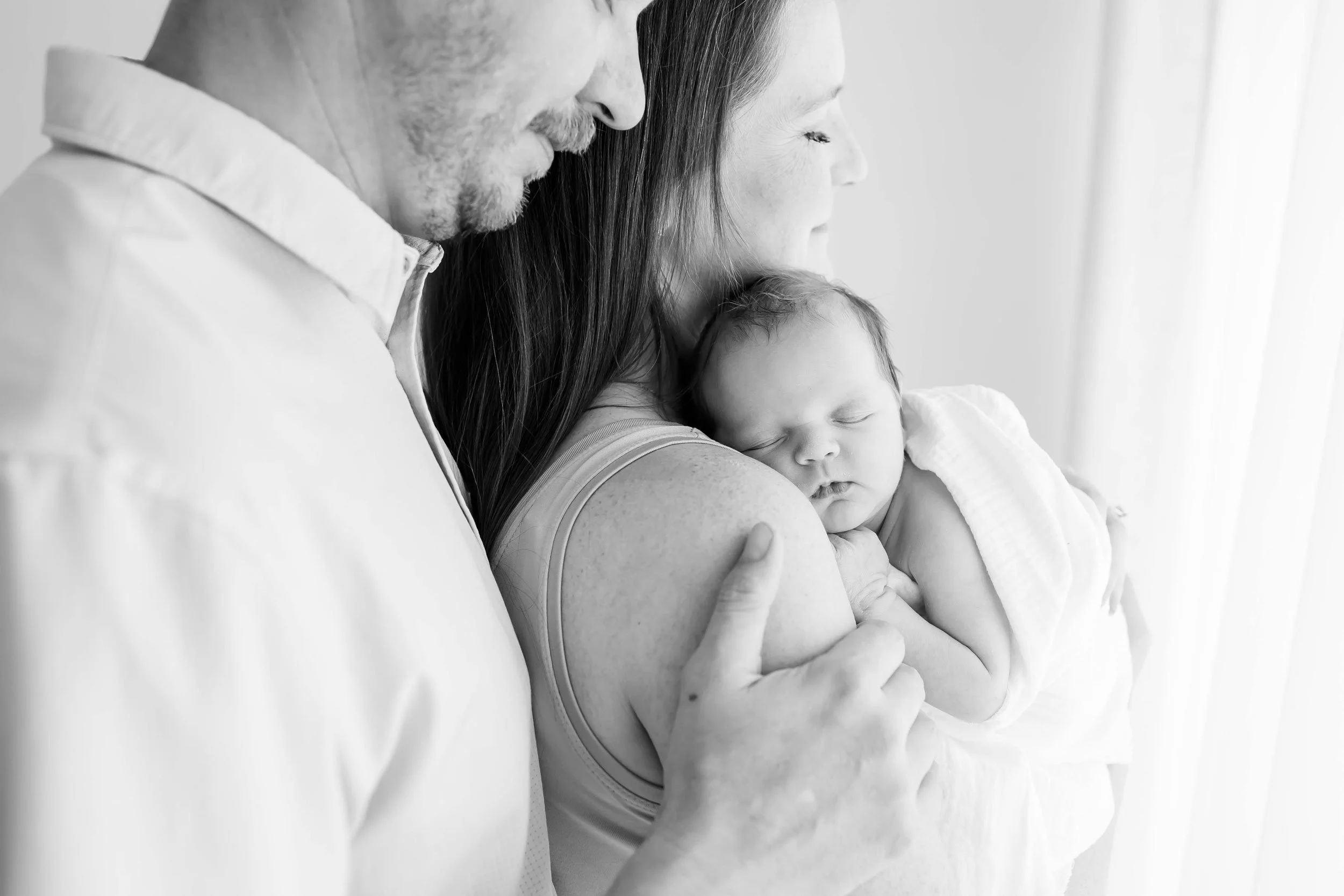 Black and white photo of a man holding a sleeping child while a woman rests her head on the mother's shoulder at East and Elm Portrait Studio in Fairport, NY, during a newborn session.
