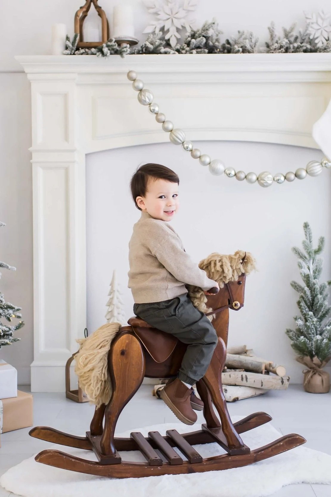 A young child smiling while riding a wooden rocking horse in a winter-themed decorated room with snowflake ornaments, small snow-covered Christmas trees, and wrapped presents during Christmas mini sessions at East and Elm Portrait Studio in Fairport,