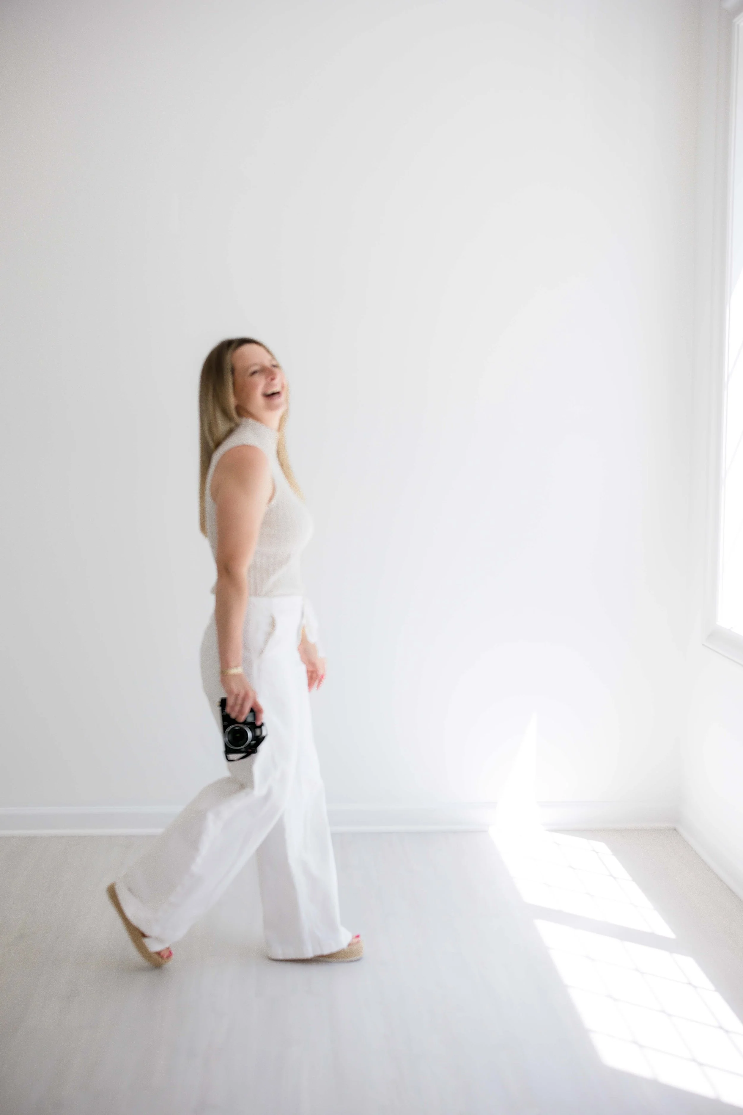 Erinn Magin, Fairport, NY family and portrait photographer, wearing a beige sleeveless top and white wide-leg pants, laughing and walking across a bright, white room with sunlight streaming through a window.