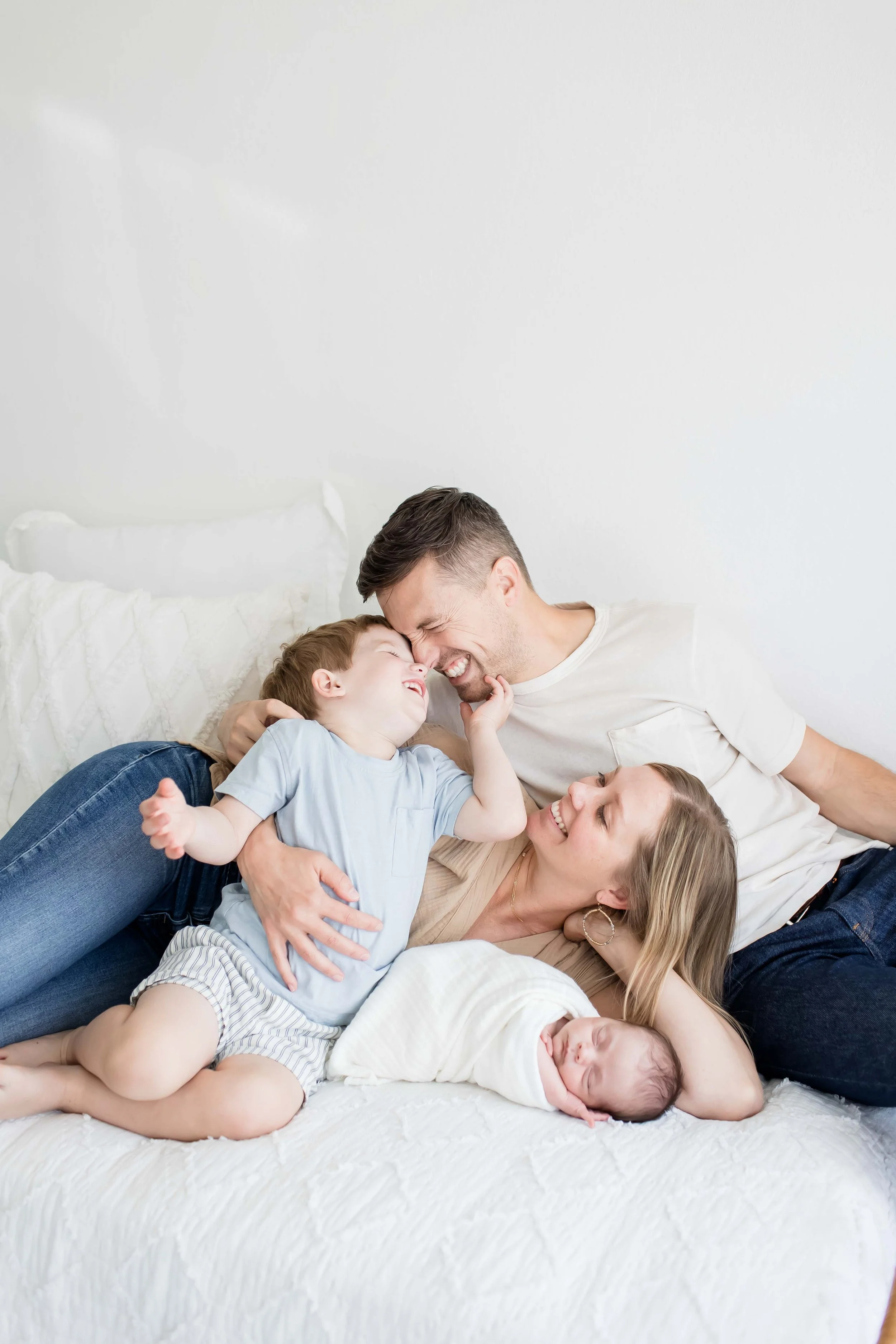 A happy family of six lying on a bed, smiling and cuddling together  at East and Elm Portrait Studio in Fairport, NY, during a family session.
