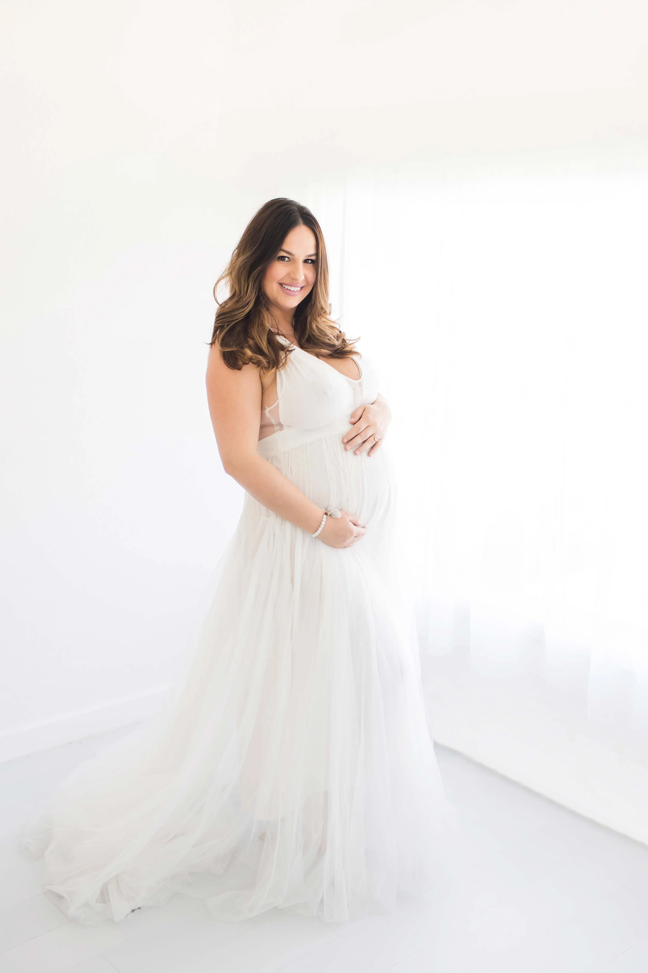 Mother wearing all white looking out window holding bump at East and Elm Portrait Studio in Fairport, NY