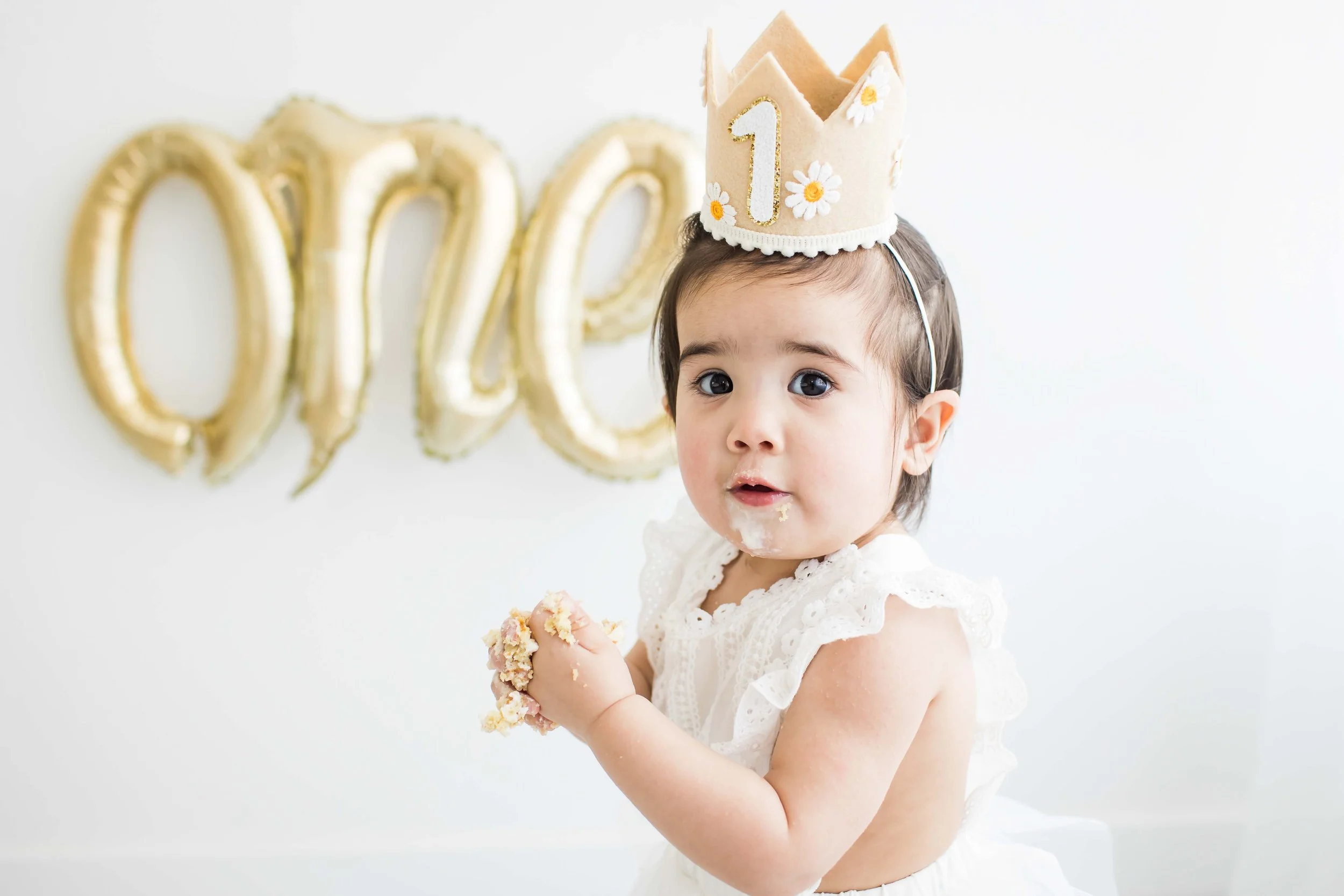 Toddler girl in a birthday crown eating cake in front of gold balloon letters spelling One, Rochester NY