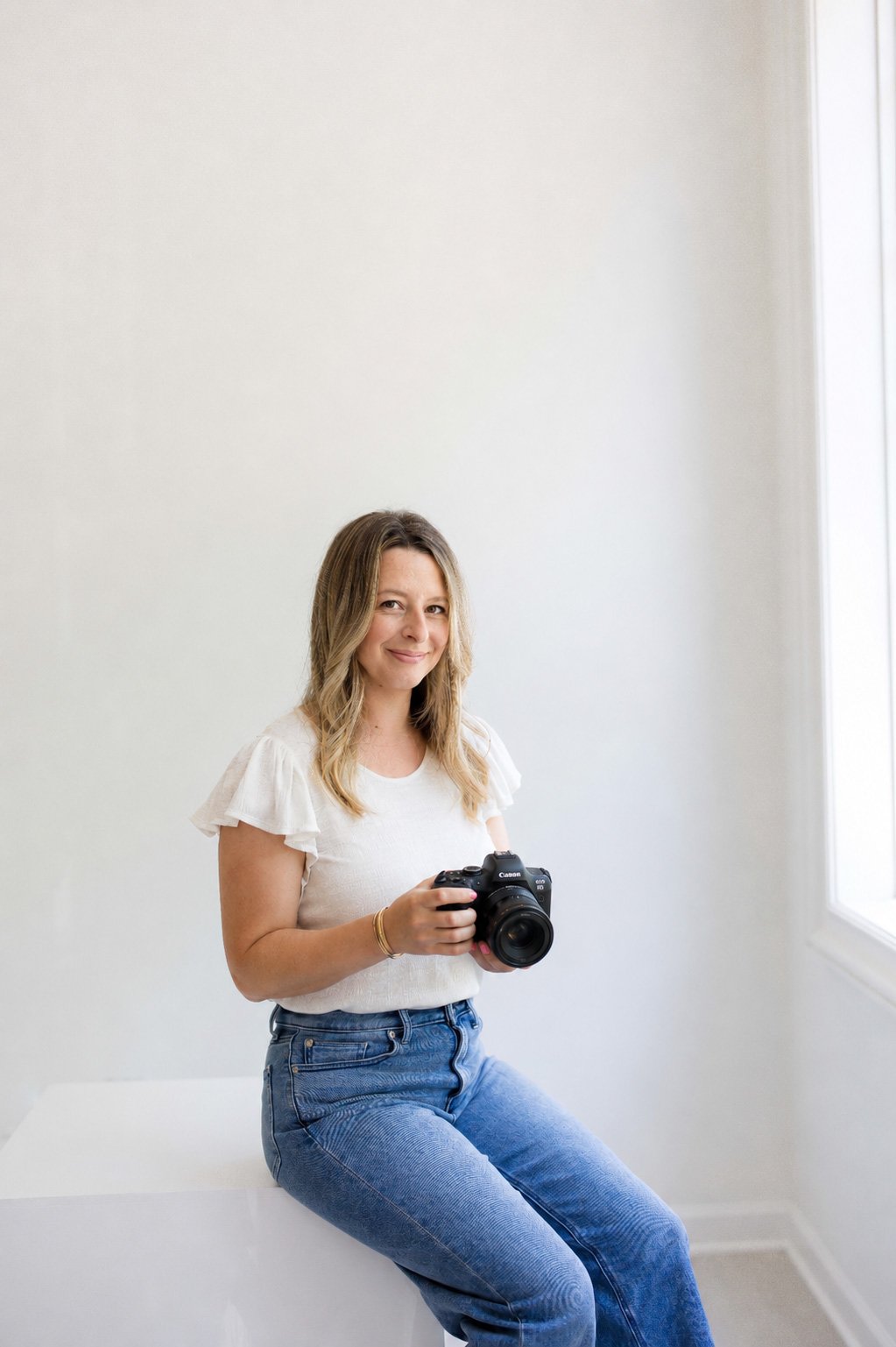 Erinn Magin, Fairport, NY family and portrait photographer, sitting on a white block, holding a Canon camera, smiling softly, wearing a white top with ruffled sleeves and blue jeans.