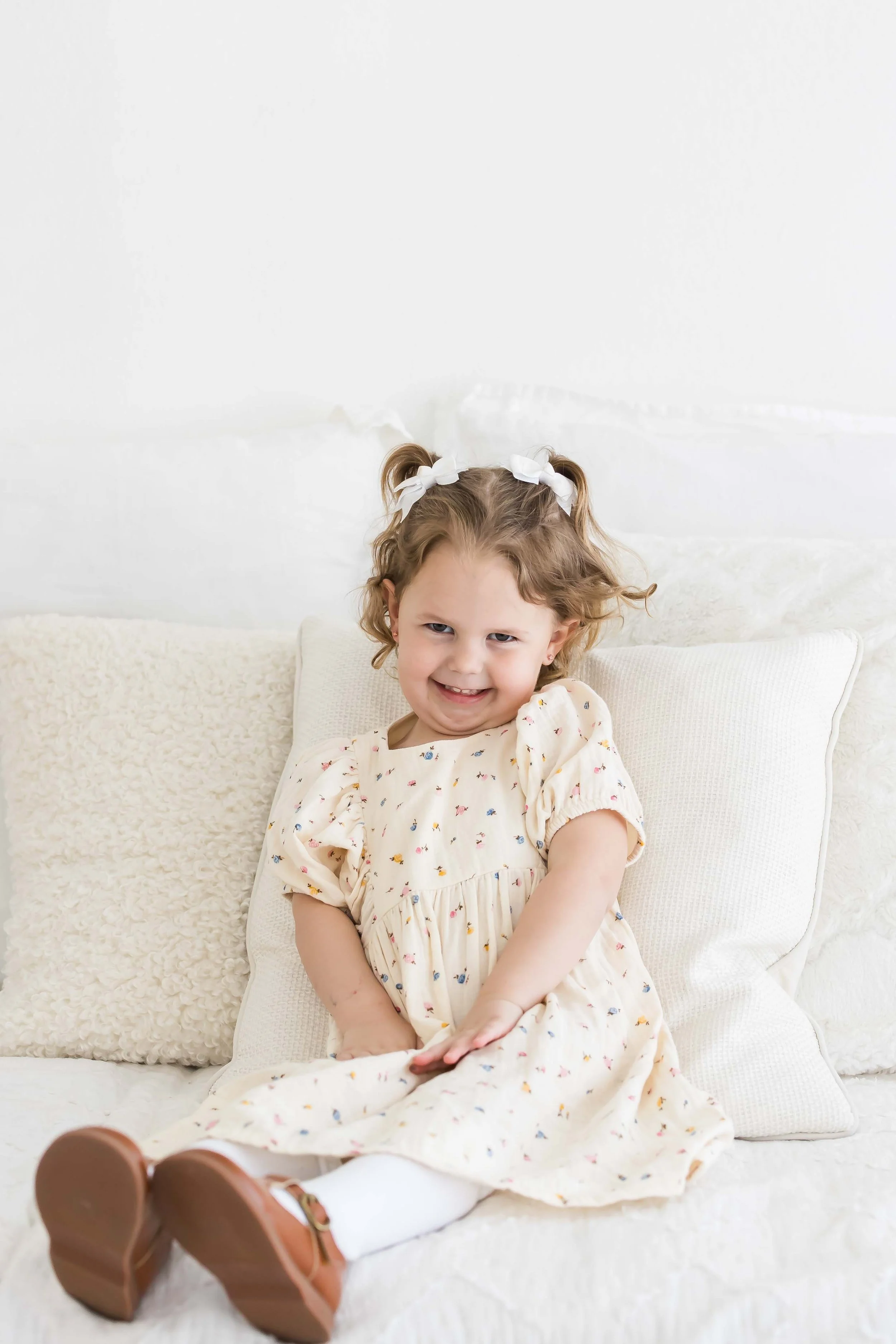 A young girl with curly hair, tied with white bows, sitting on a beige textured blanket and pillows, smiling at the camera at East and Elm Portrait Studio in Fairport, NY, during a family session.