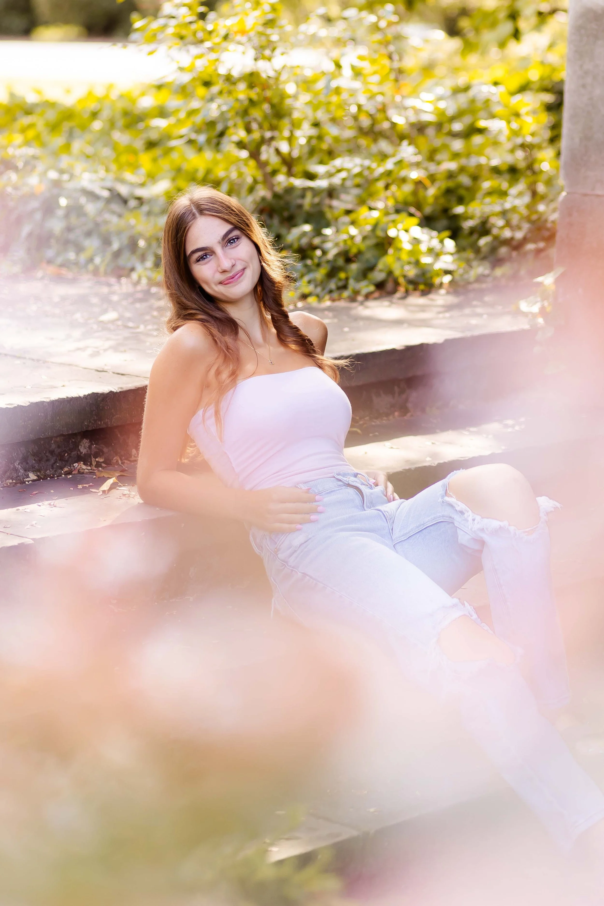 A young woman with long wavy brown hair and a white tank top sitting on a stone ledge outside, smiling at the camera with greenery in the background during her senior portrait session with East and Elm Portrait Studio in Rochester, NY.
