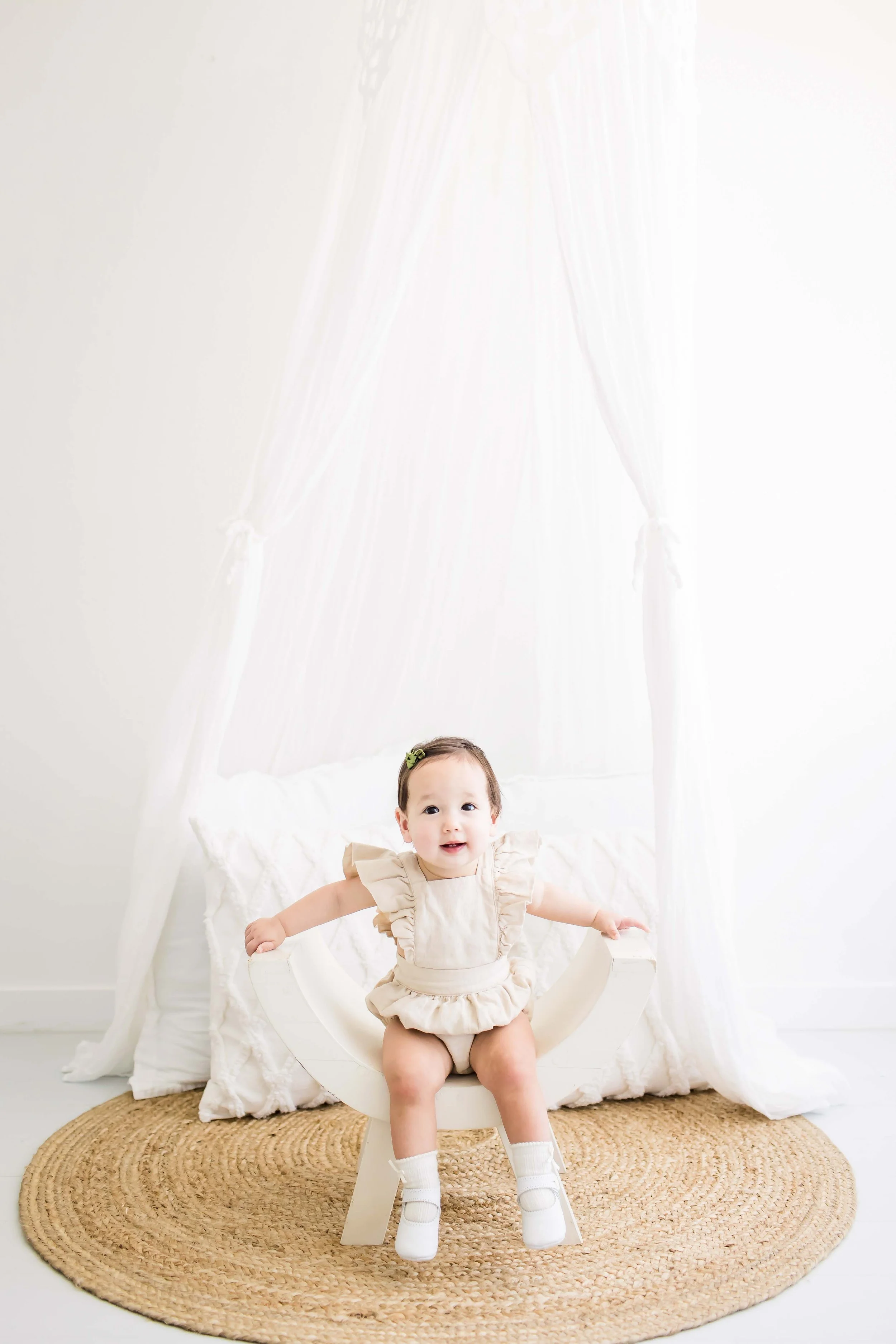 A baby sitting on a small white chair in front of a white bed with a canopy, on a round jute rug, looking at the camera with arms extended at East and Elm Portrait Studio in Fairport, NY, during a milestone session.