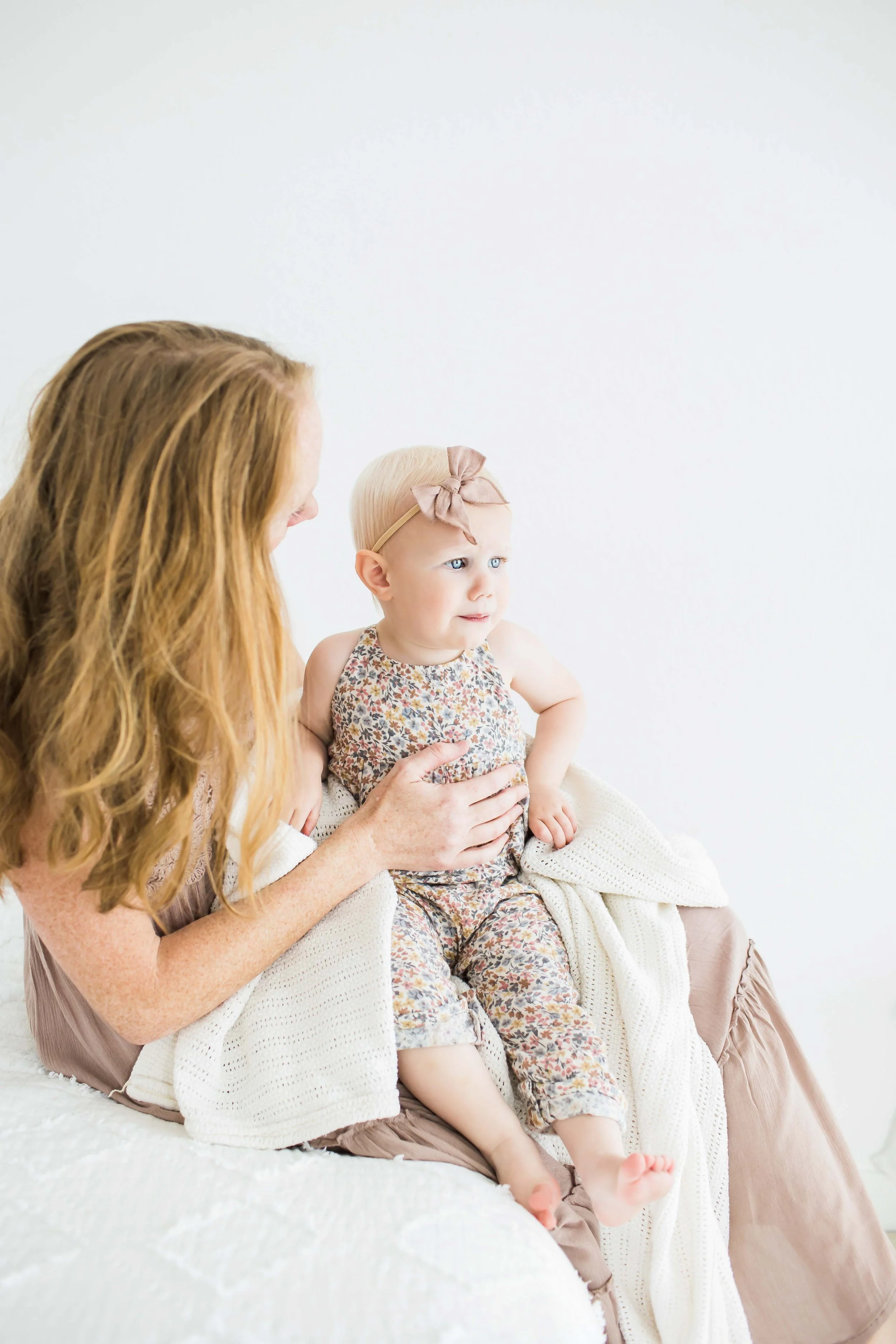A woman holding a toddler girl with blonde hair sitting on a bed with a white blanket and beige cushions at East and Elm Portrait Studio in Fairport, NY, during a family session.