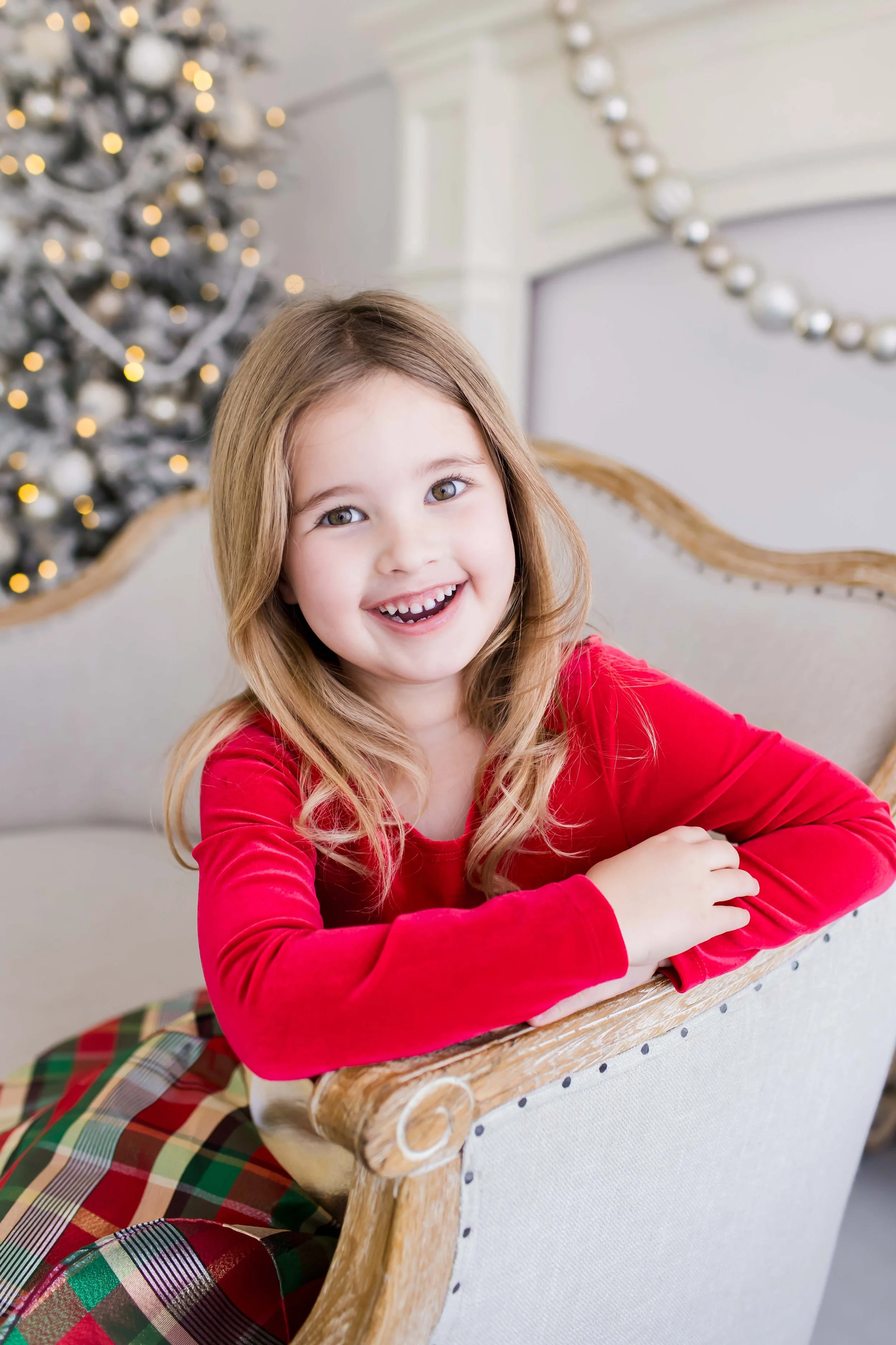 A smiling young girl with brown hair, wearing a red top and plaid skirt, sitting on a beige upholstered chair in front of a decorated Christmas tree with lights at East and Elm Portrait Studio in Fairport, NY, during a christmas mini session.