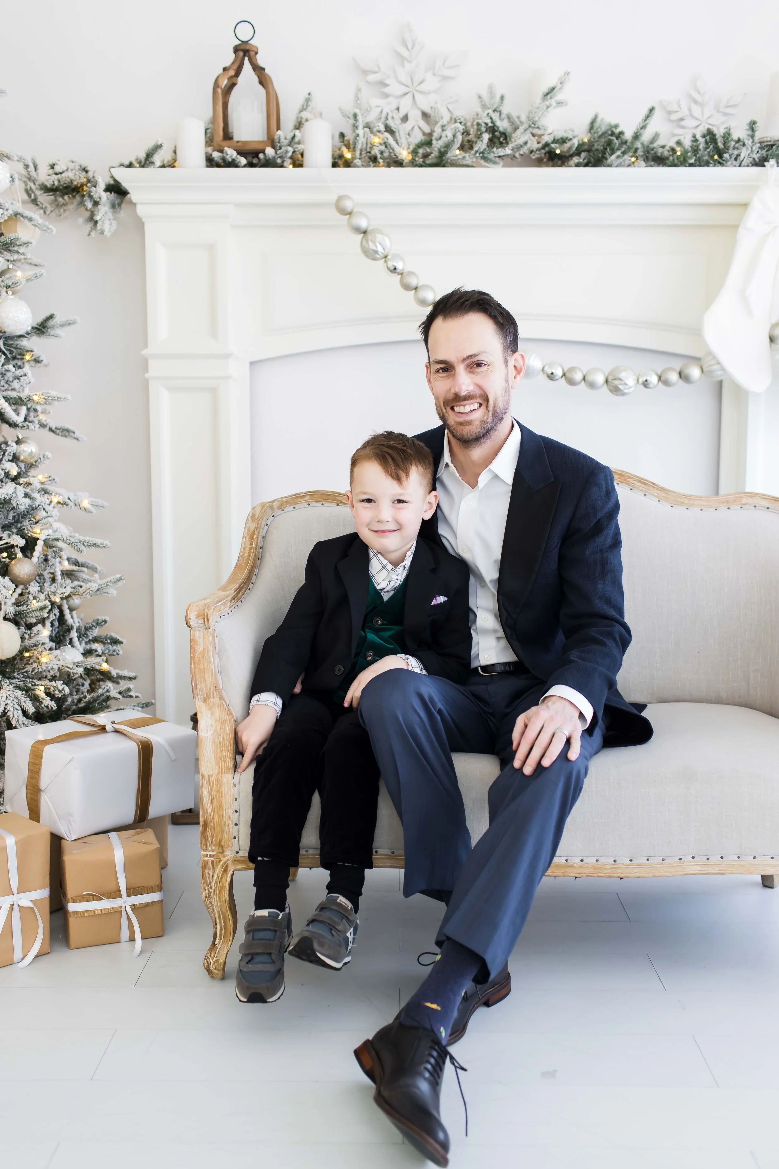 Dad and son sitting on a vintage-style sofa in a festively decorated living room with Christmas tree and wrapped gifts, smiling at the camera during Christmas mini sessions at East and Elm Portrait Studio in Fairport, NY