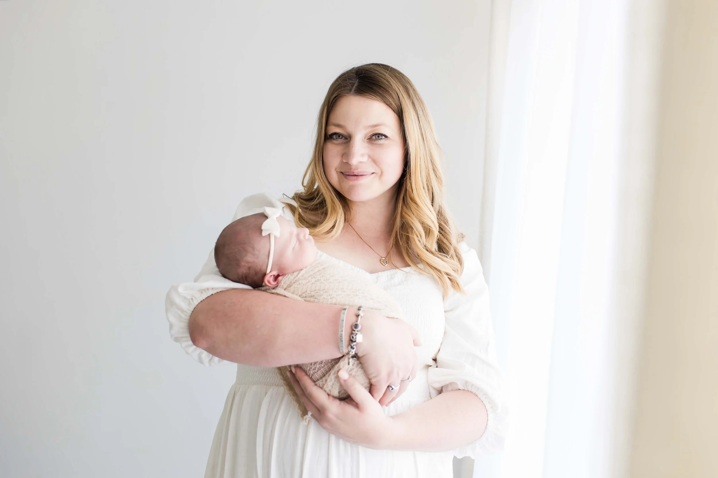 A mom holding a newborn baby wrapped in a beige blanket. The woman is smiling and wearing a white dress from the client closet at East and Elm Portrait Studio in Fairport, NY, during a newborn session.
