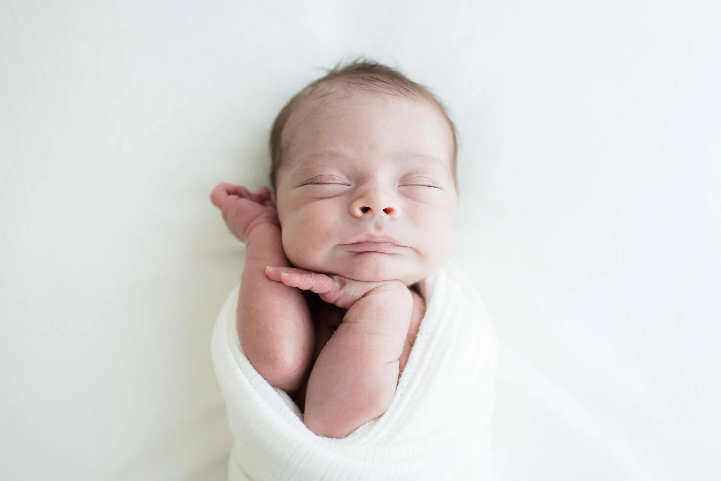 Close-up of a sleeping newborn baby with a hand gently touching their face, against a plain white background at East and Elm Portrait Studio in Fairport, NY, during a newborn session.