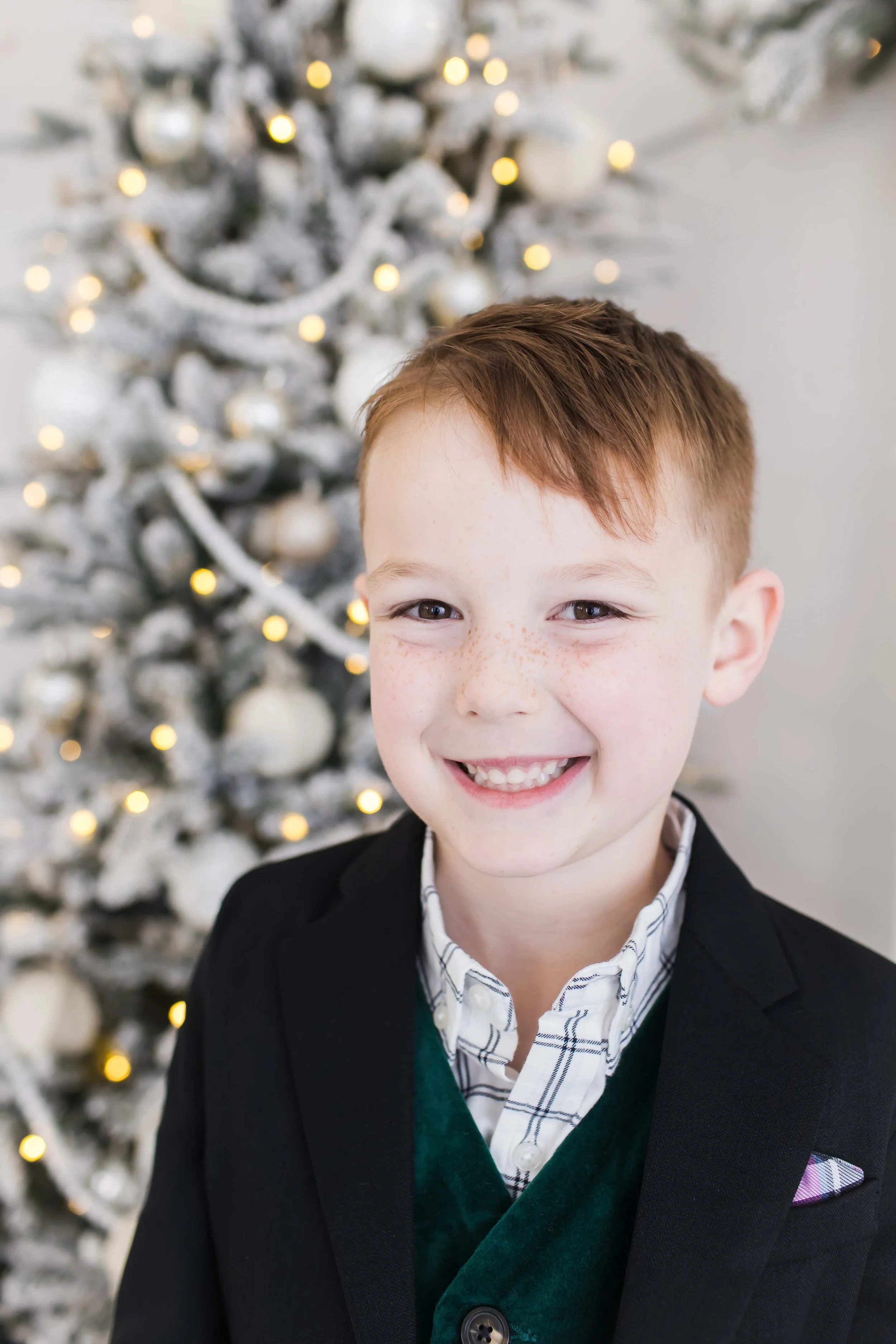 A young boy with short brown hair, freckles, and a big smile standing in front of a decorated Christmas tree with white ornaments and warm lights during Christmas mini sessions at East and Elm Portrait Studio in Fairport, NY