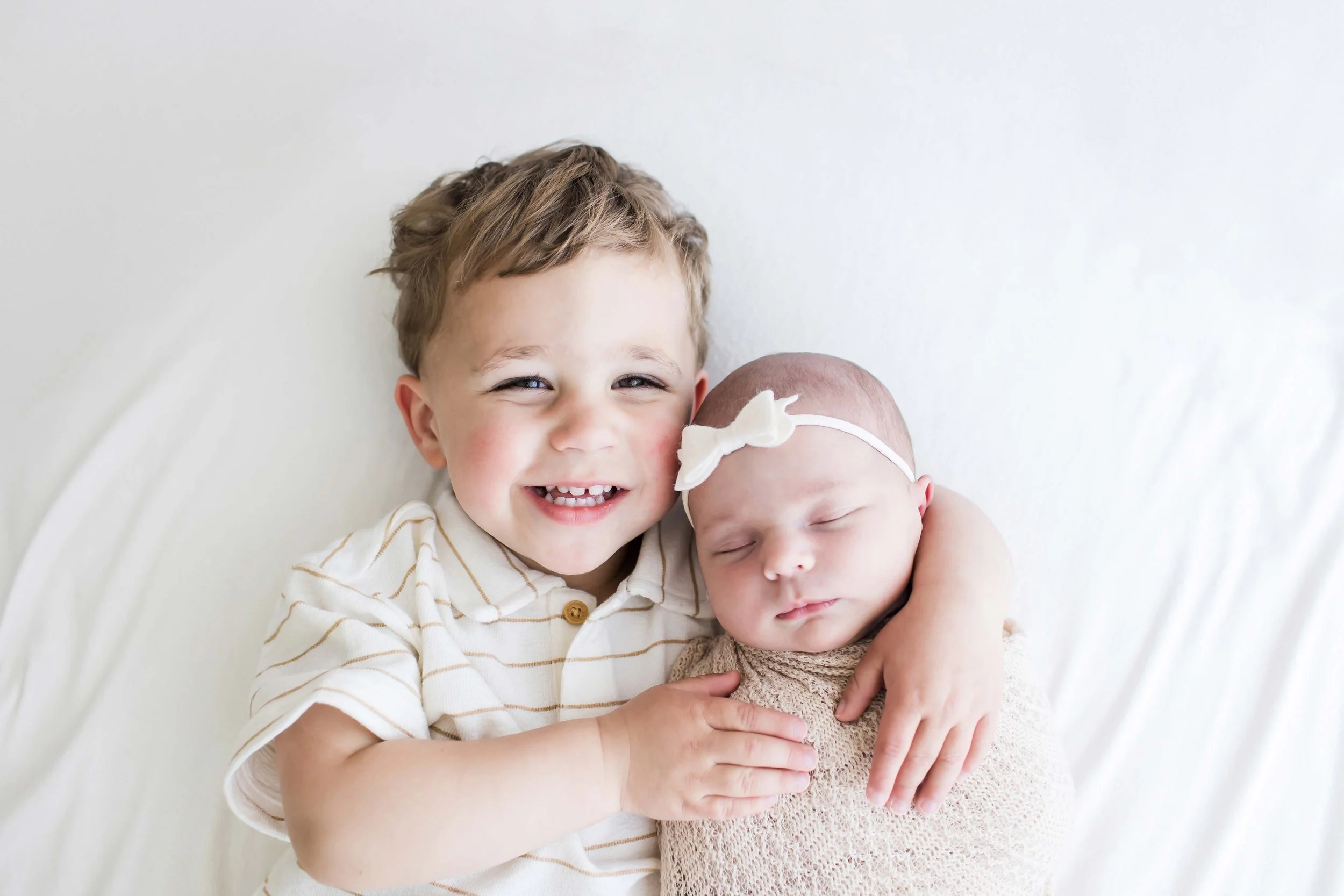 A young boy smiling and hugging a sleeping newborn baby girl at East and Elm Portrait studio in Fairport, NY during newborn session.