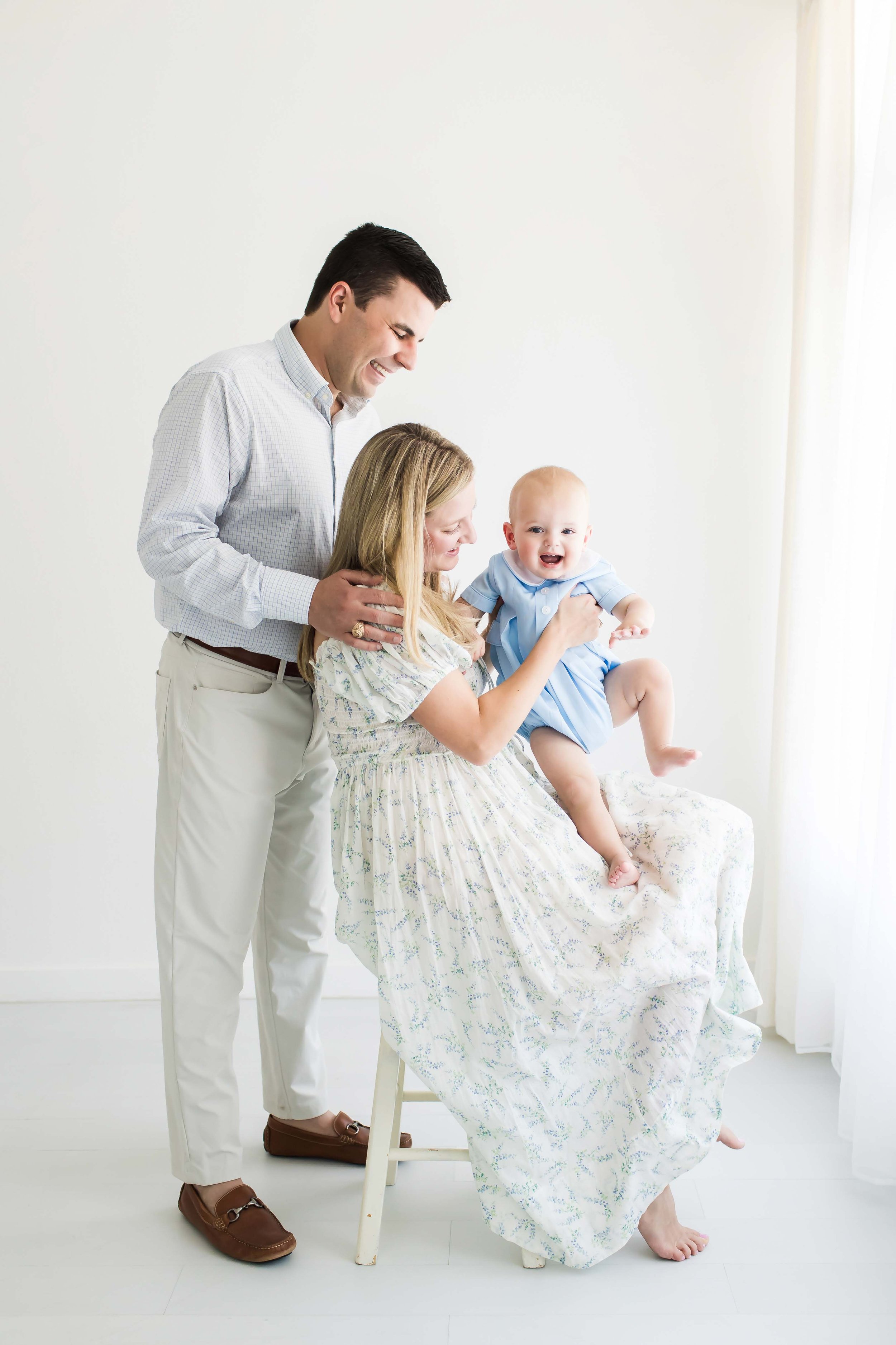 A family of three celebrating a baby in a bright, white room at East and Elm Portrait Studio in Fairport, NY, during a family session.
