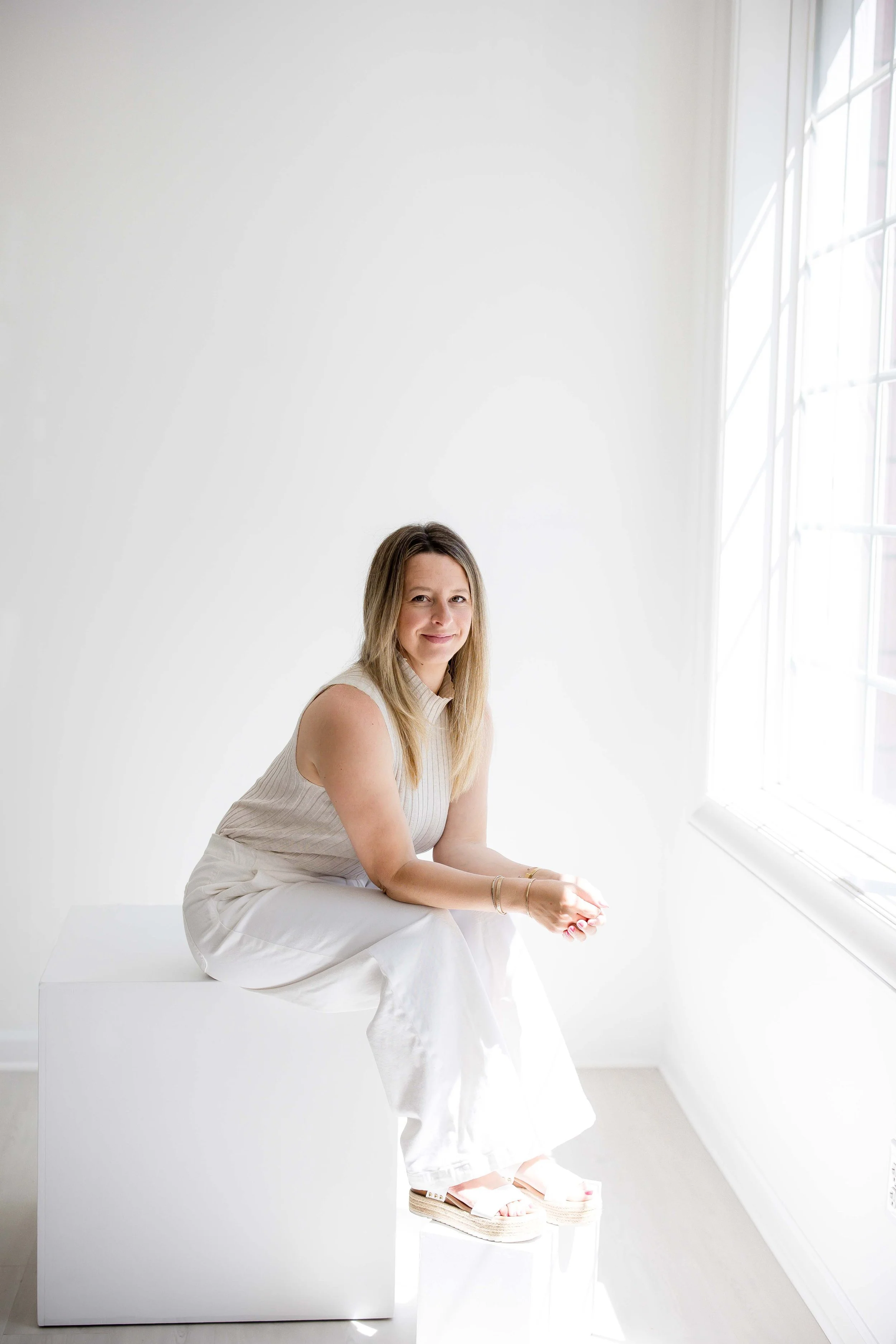 Erinn Magin, owner of East and Elm Portrait studio, sitting on a white cube near a large window with sunlight streaming in, wearing white pants and a light-colored sleeveless top, smiling and looking at the camera.