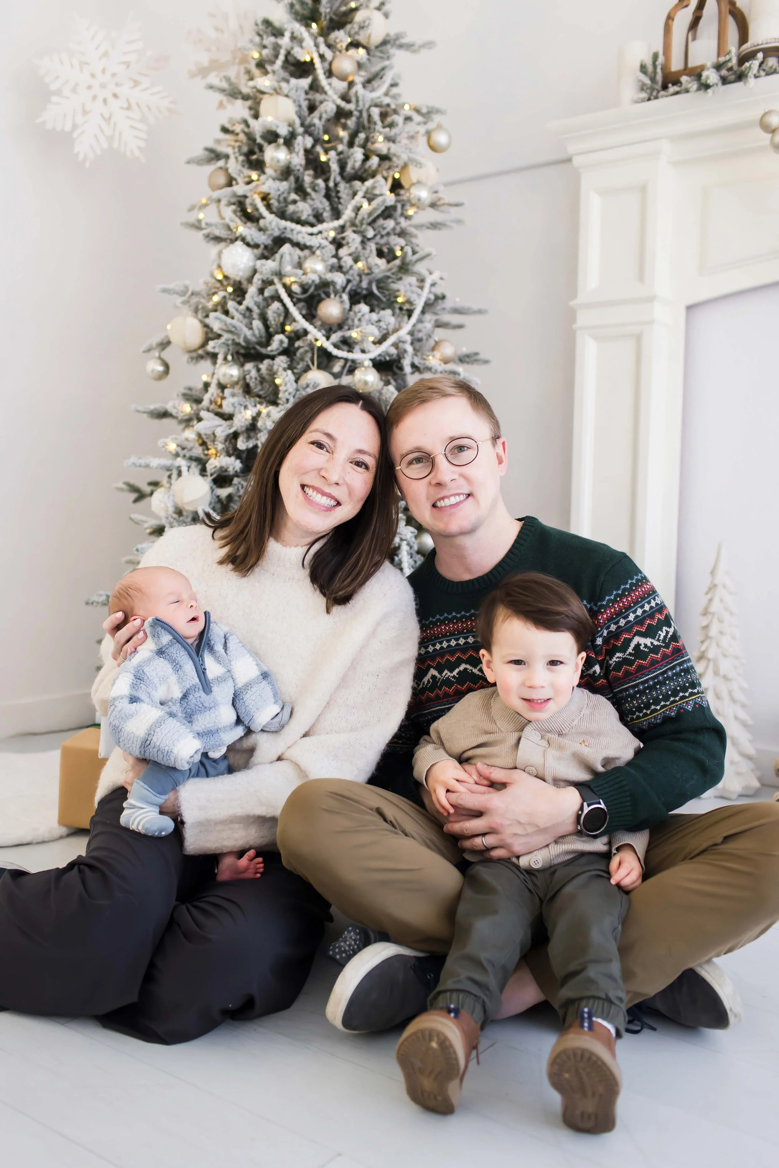 A family of four with a baby in front of a decorated Christmas tree with ornaments and lights during Christmas mini sessions at East and Elm Portrait Studio in Fairport, NY
