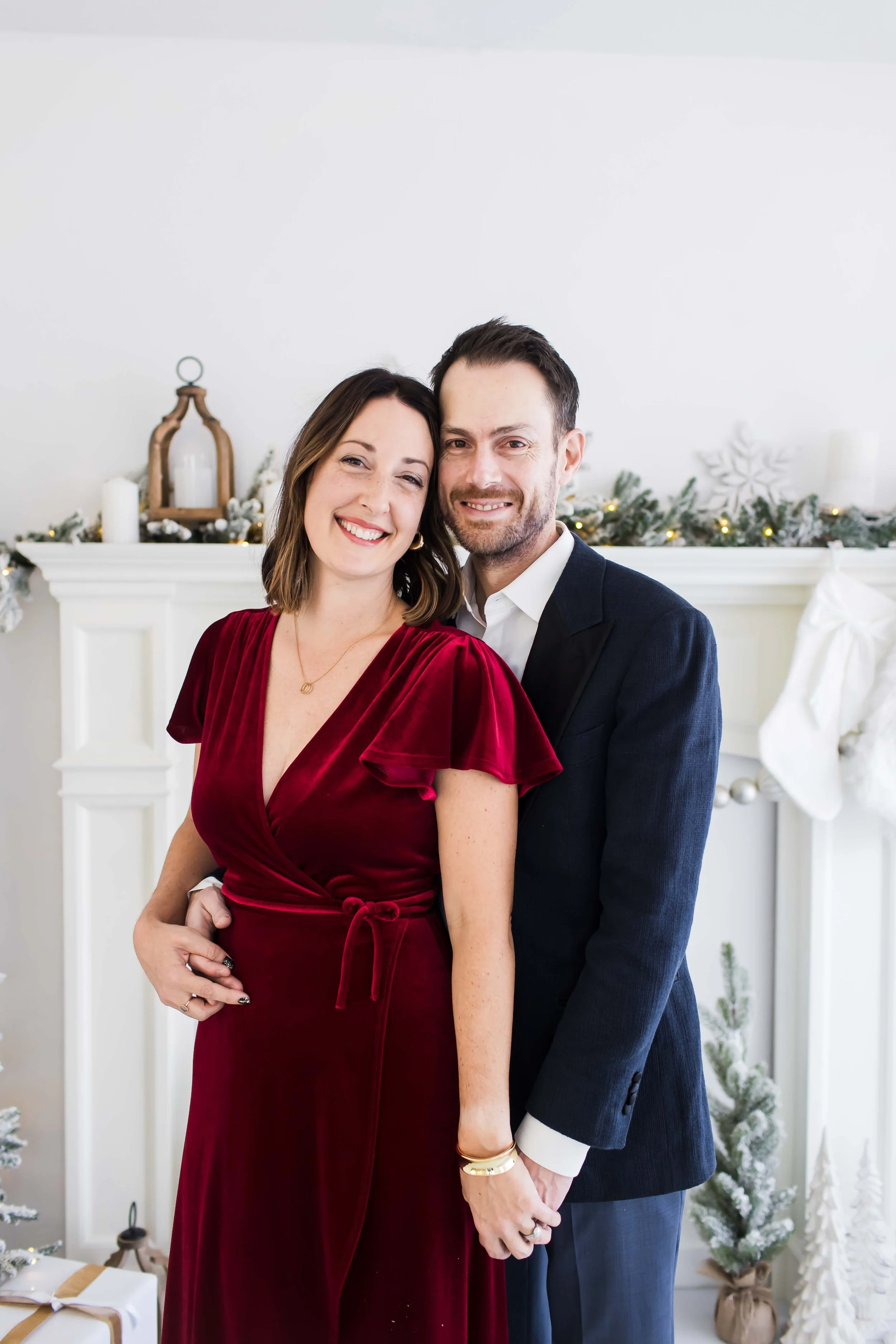 A smiling couple standing in front of a decorated fireplace with greenery, candles, and a large snowflake ornament at East and Elm Portrait Studio in Fairport, NY, during a Christmas mini session.