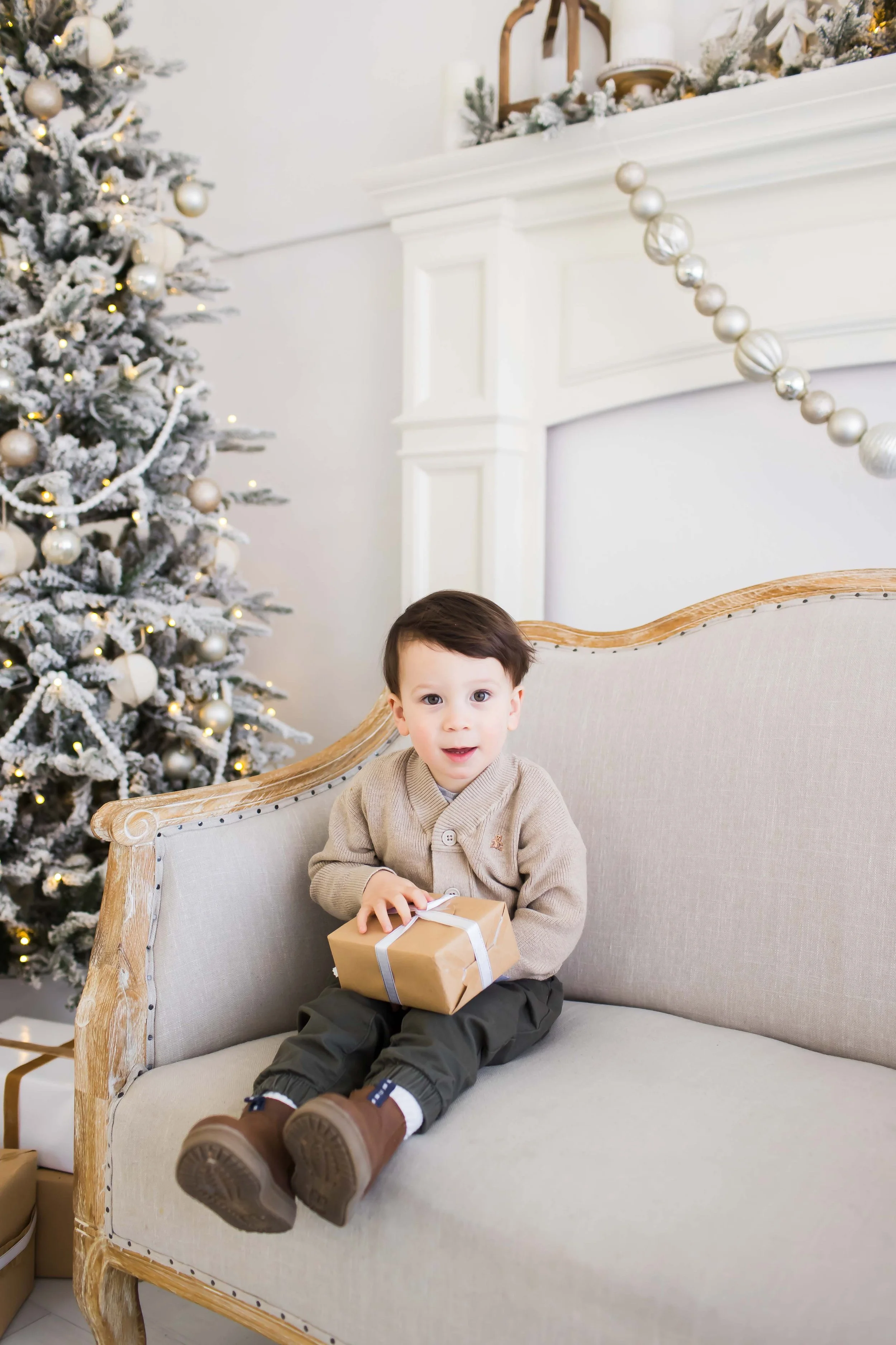 A young boy sitting on a beige upholstered sofa, holding a wrapped gift box, in front of a white Christmas tree at East and Elm Portrait Studio in Fairport, NY, during a Christmas mini session.