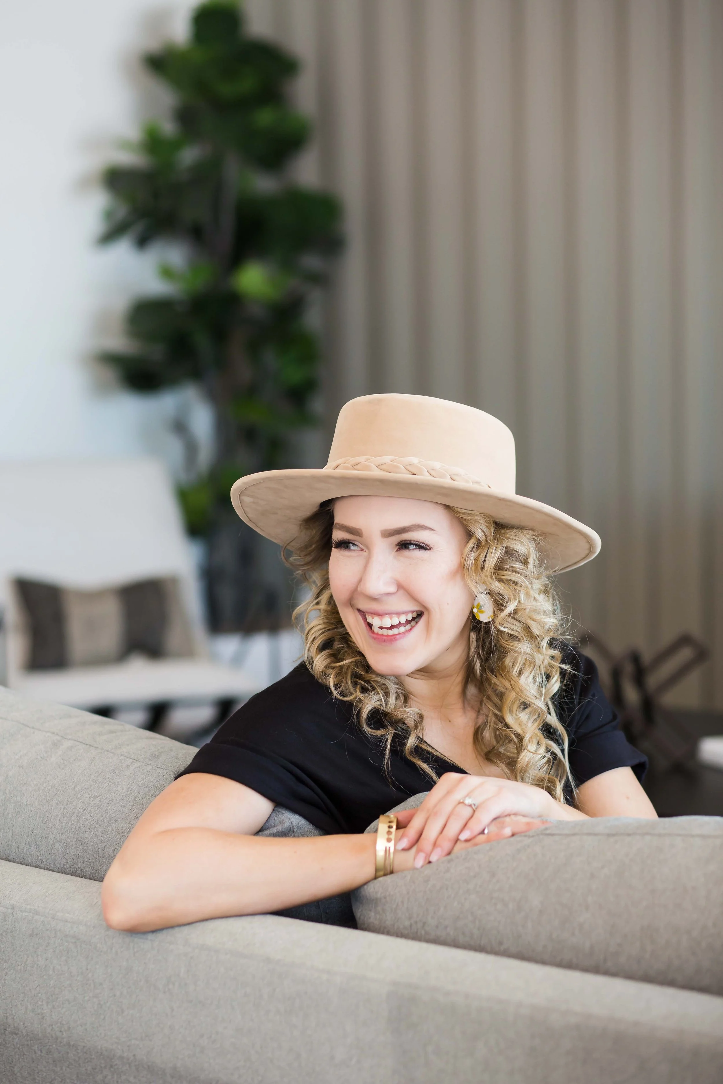 A woman with curly blonde hair, wearing a beige hat and a black top, sitting on a light gray couch in a modern, well-lit room, smiling and looking to her left during a branding and headshot session for business owners in Rochester, NY with East and E