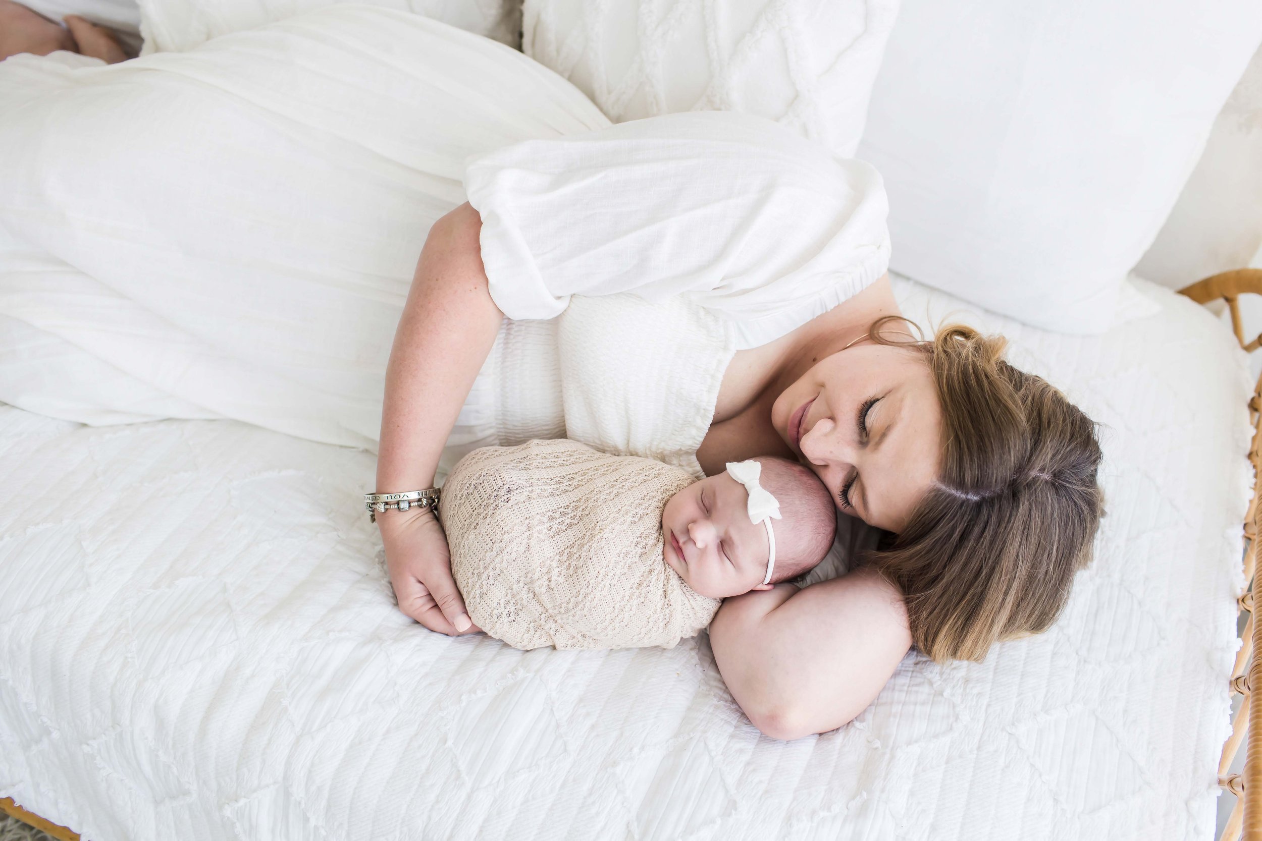 A woman with brown hair lying on a bed, close to a sleeping newborn baby wrapped in a beige blanket, both wearing items from the client closet at East and Elm Portrait Studio in Fairport, NY, during a newborn session.