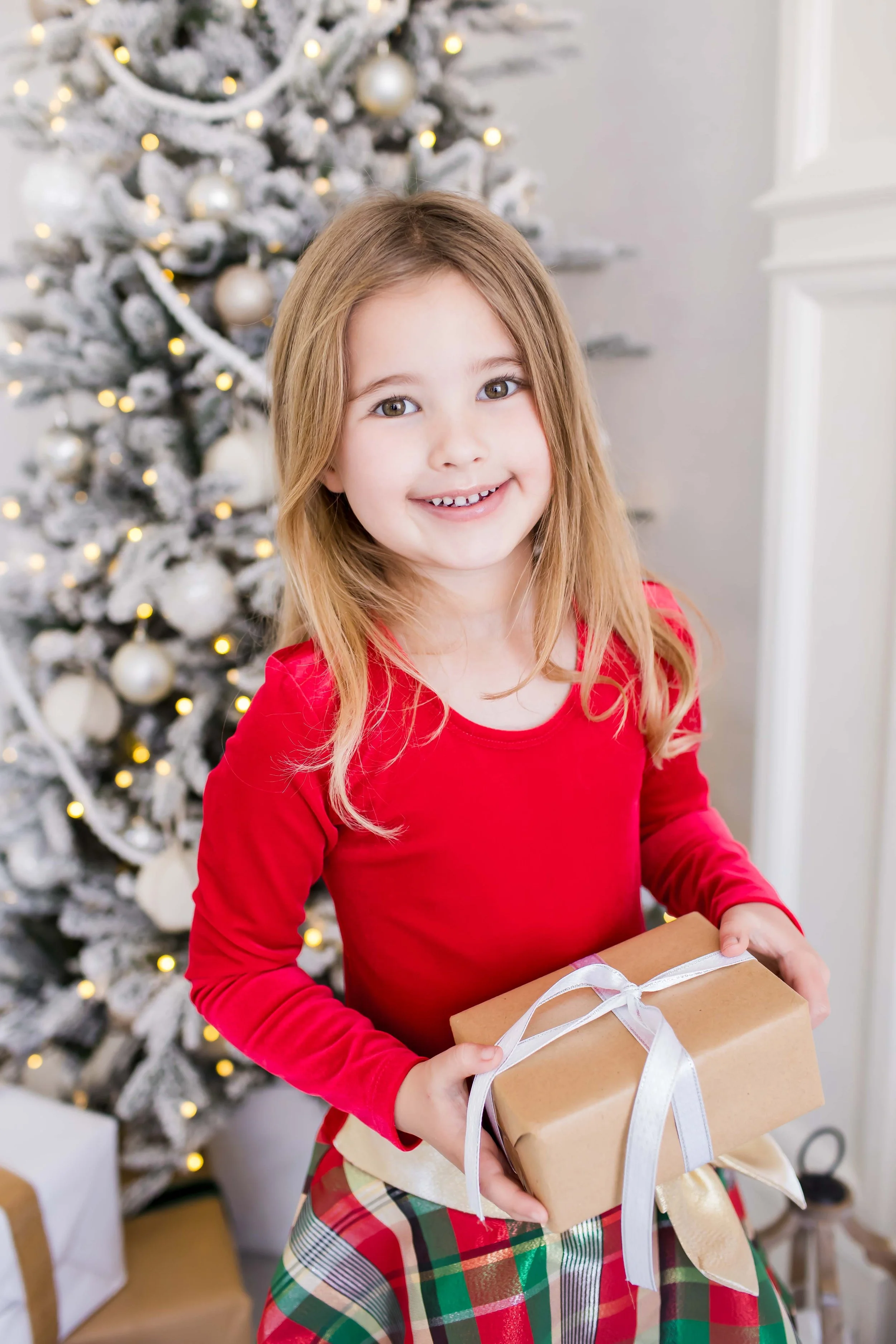 A young girl in a Christmas dress holding a wrapped gift with a white ribbon in front of a decorated Christmas tree with ornaments and lights  at East and Elm Portrait Studio in Fairport, NY, during a Christmas mini session.
